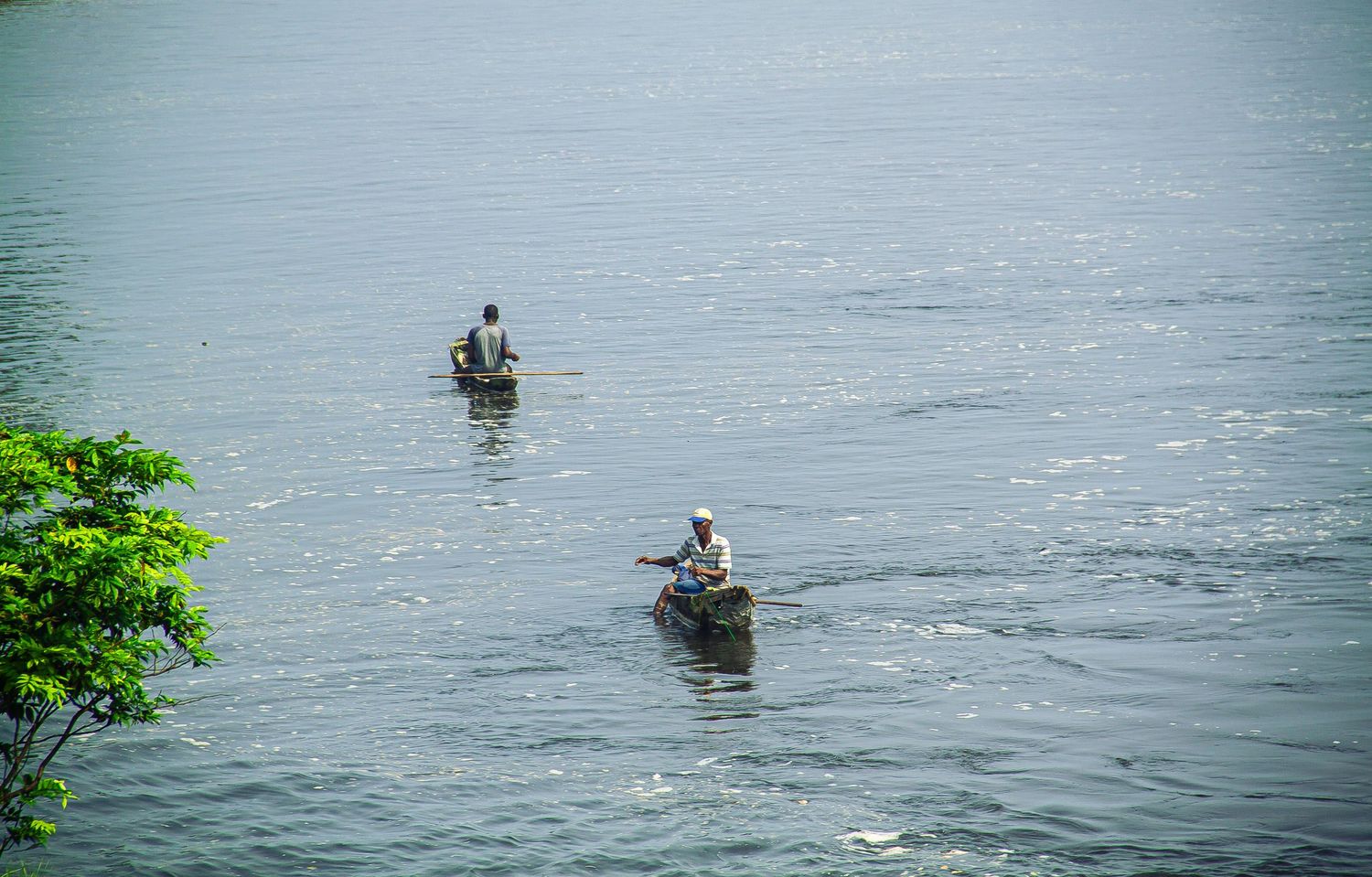 Two fishermen paddling wooden canoes across a calm grey river lined with tropical greenery.