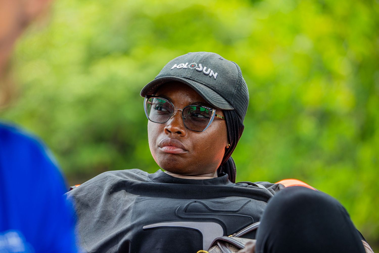 Person wearing black cap and sunglasses relaxing outdoors with green trees behind them.