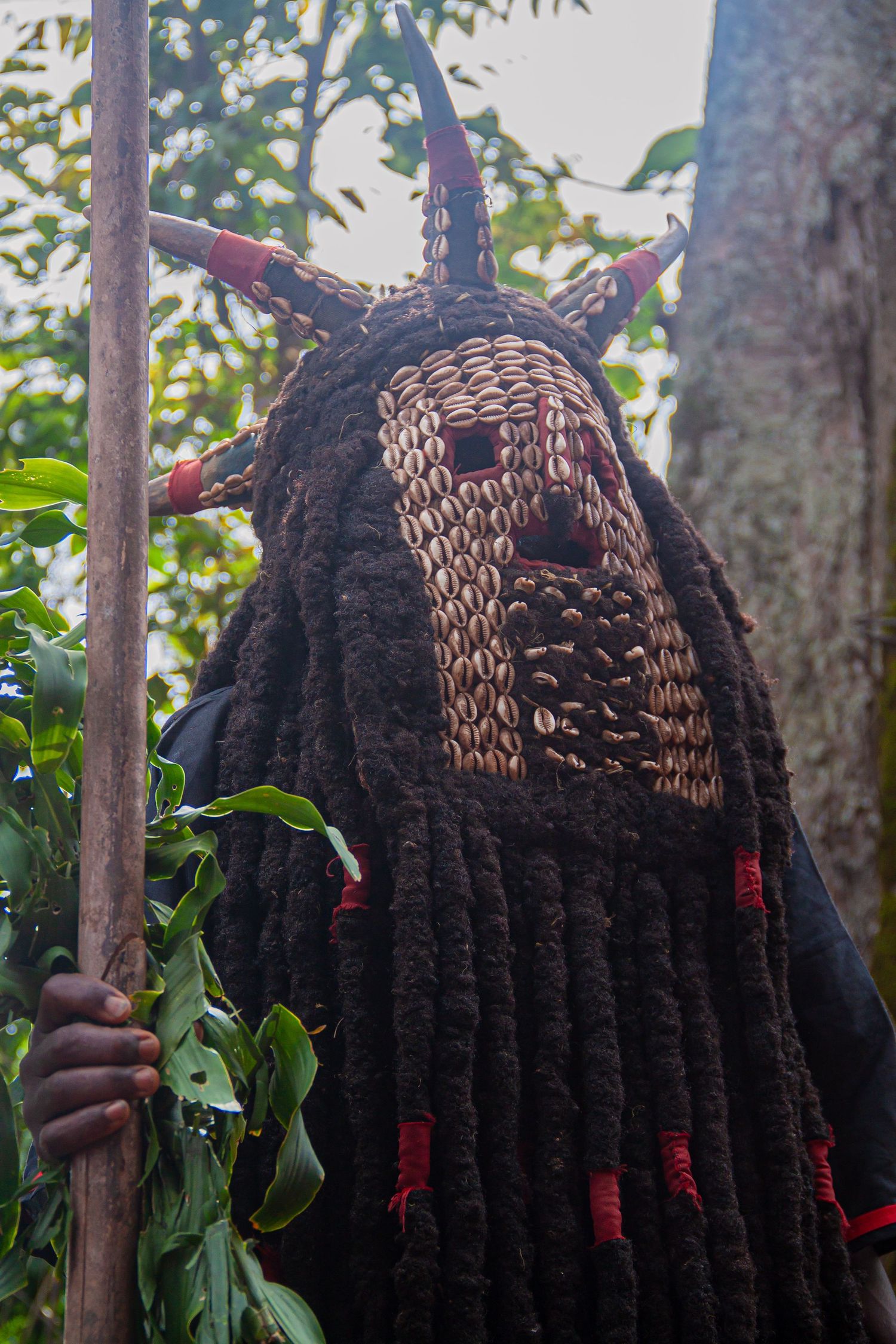 A traditional African masquerade costume featuring cowrie shell mask, long dreadlocks, and horns held by a performer.