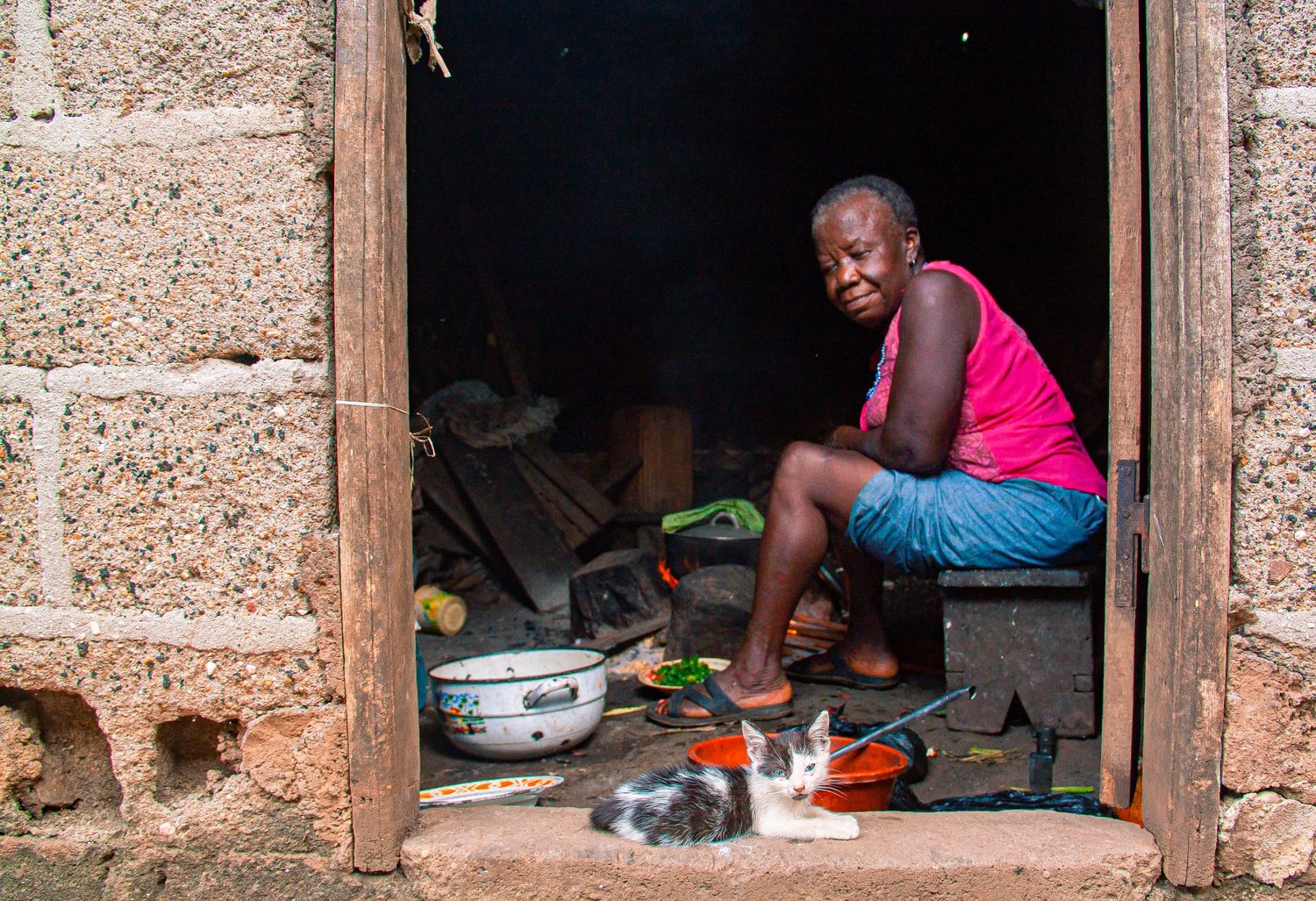 A young boy sits in a doorway of a stone building with a cat resting nearby and cooking items around him.