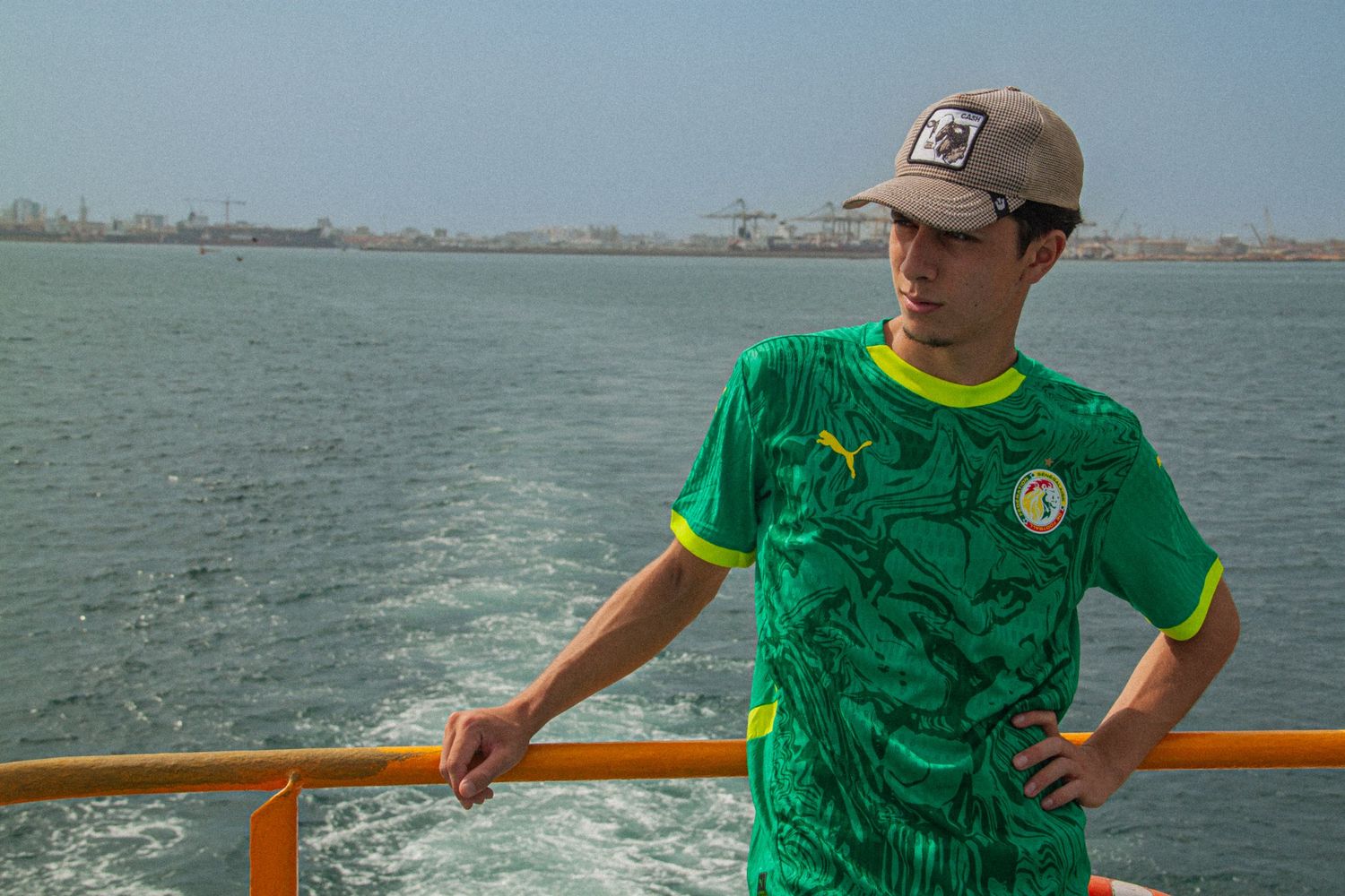 A person in a green Senegal Puma jersey stands on a ferry deck overlooking the ocean on a sunny day.