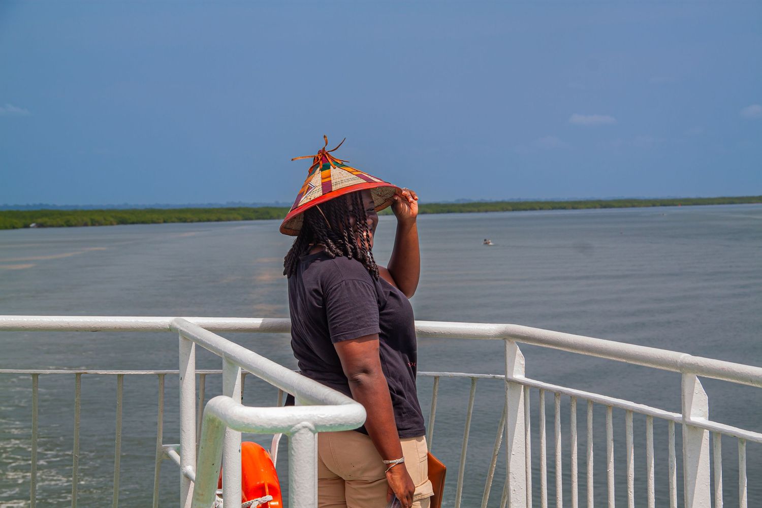 Person wearing a straw hat standing on a boat deck overlooking a vast open waterway.