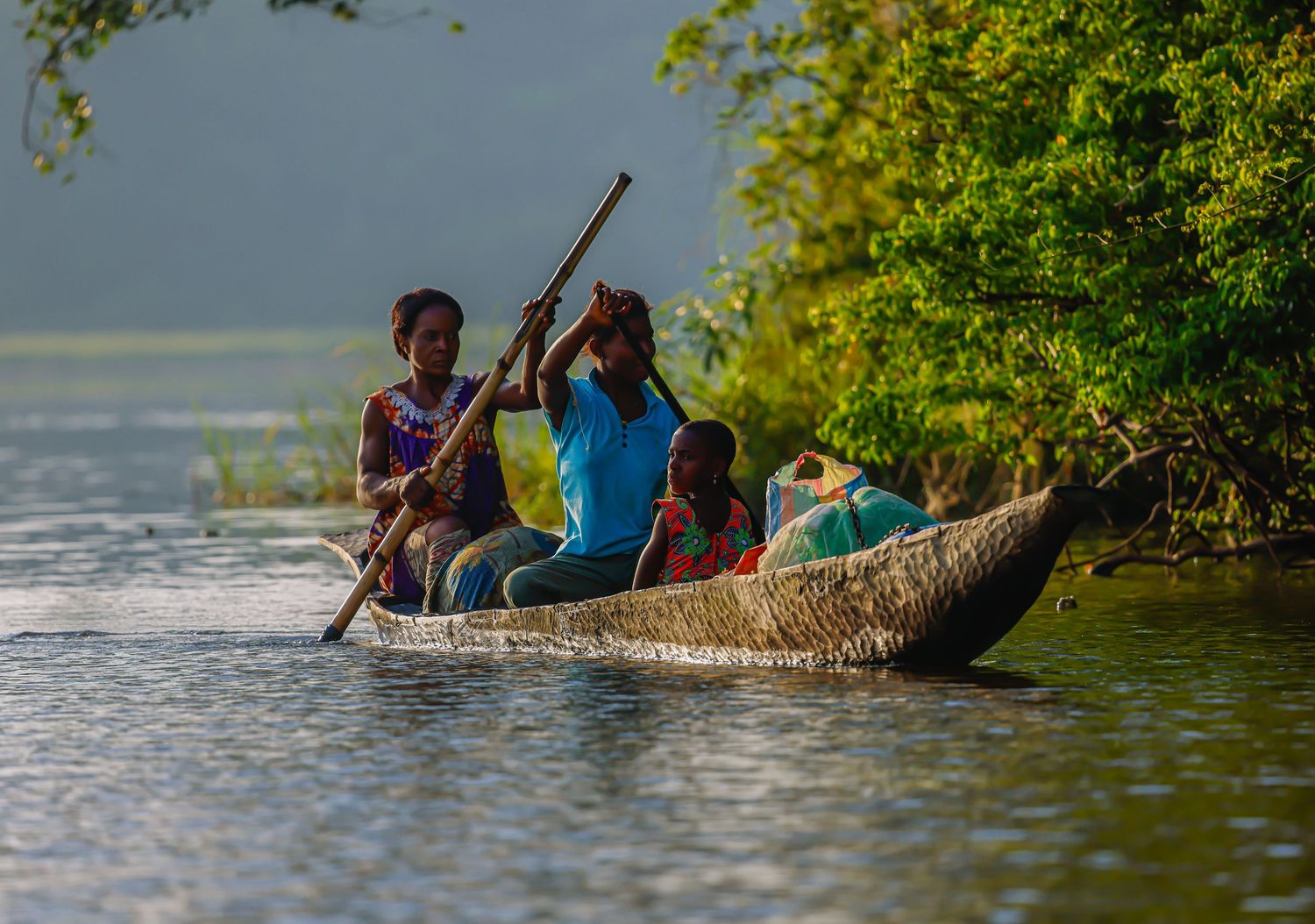 Children and adults paddling a traditional reed canoe on a calm river surrounded by green trees.