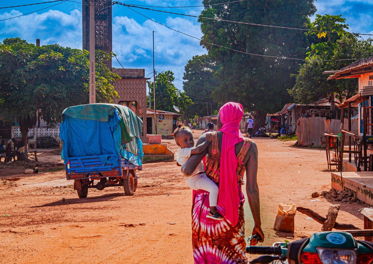 Woman in pink wrap carrying baby walks along dusty African village road with tuk-tuk passing by.