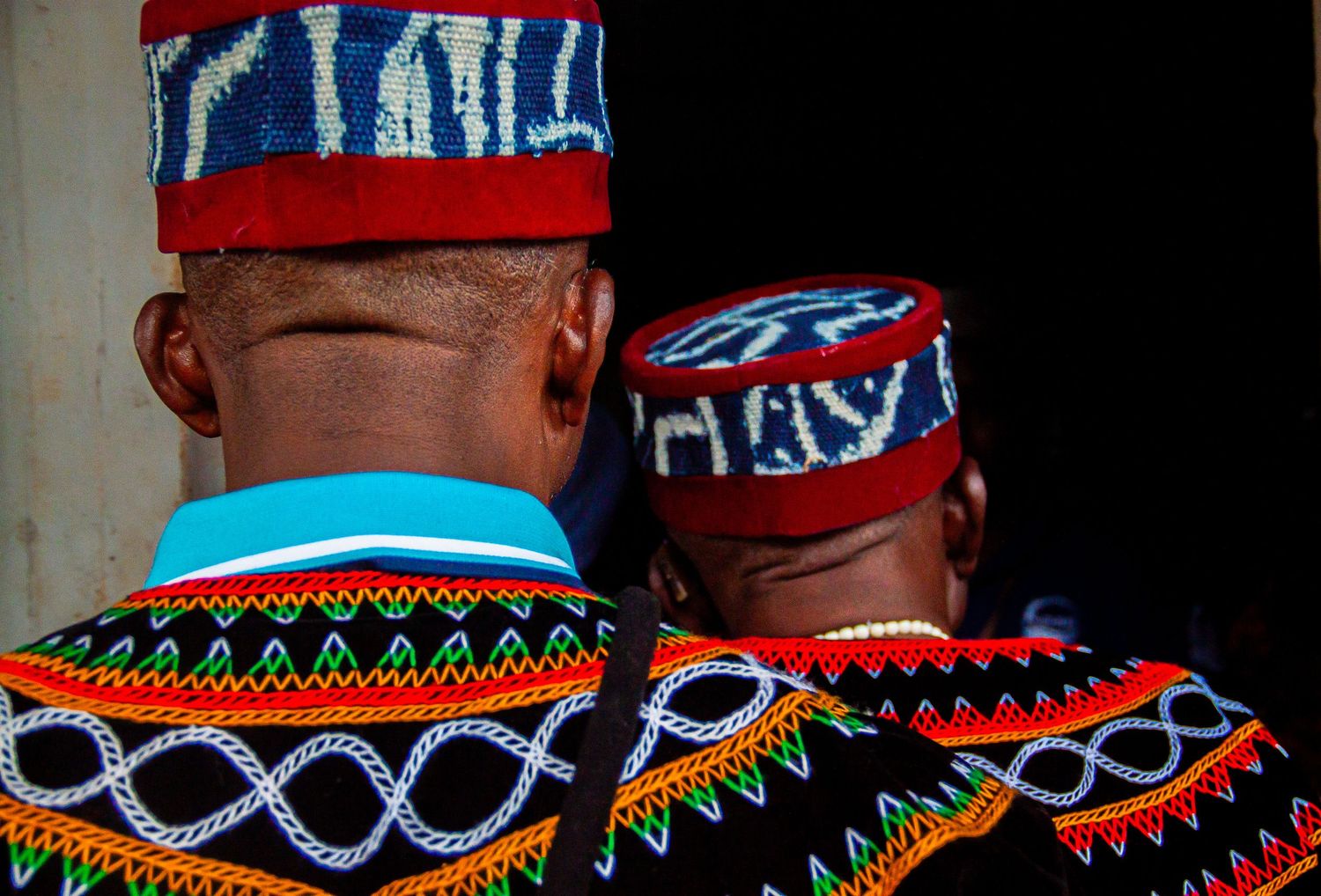 Close-up rear view of two men wearing traditional Cameroonian embroidered caps and Ndop fabric clothing.