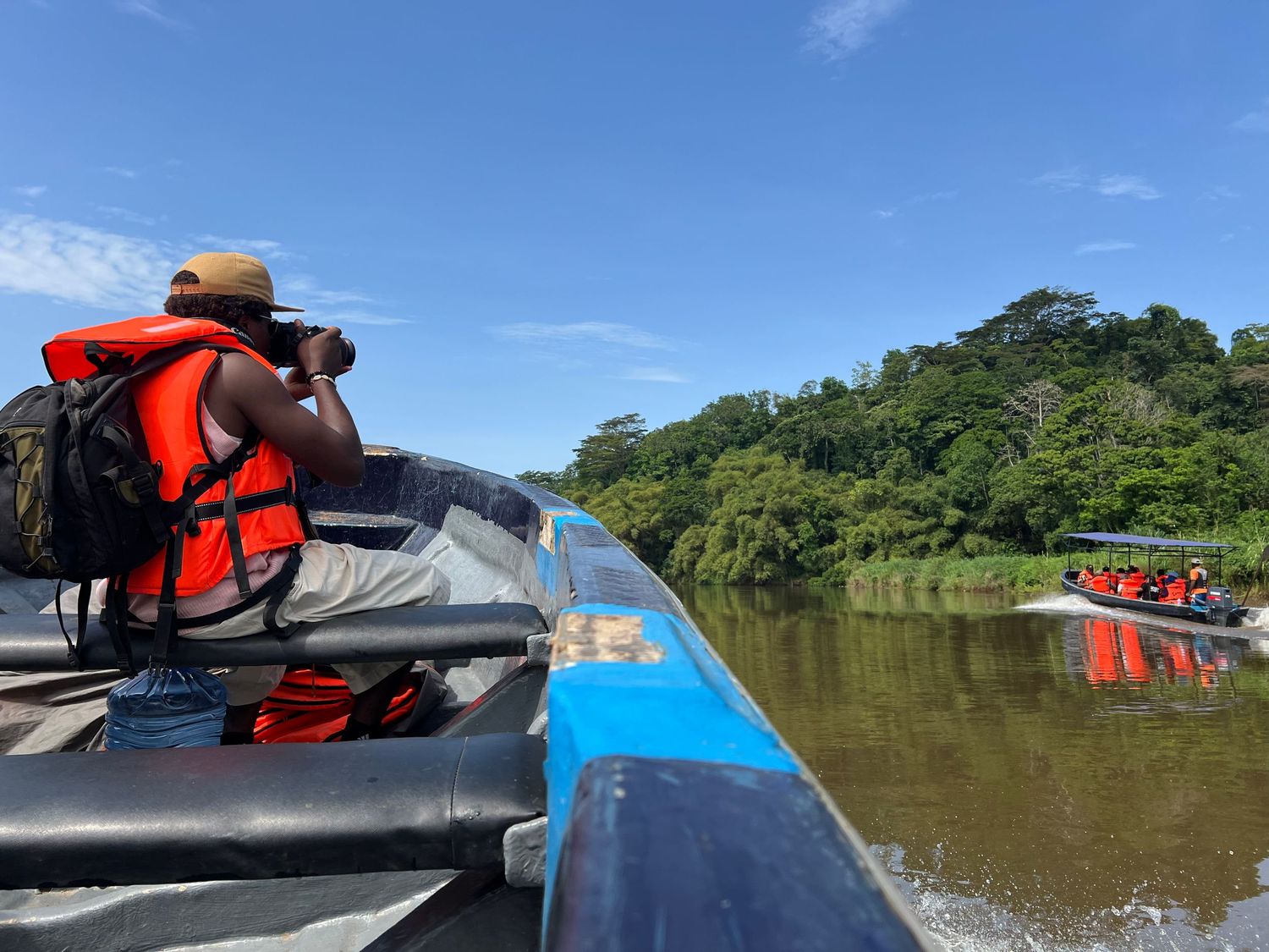 A photographer uses binoculars on a boat traveling along a tropical river surrounded by jungle.