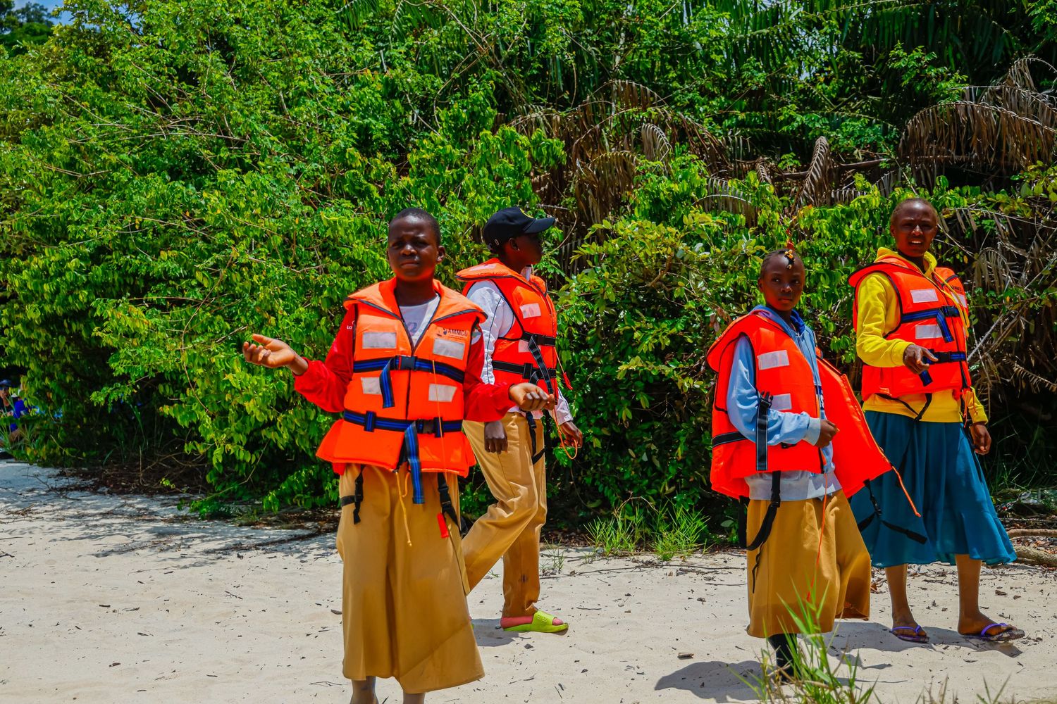 Four people wearing orange life jackets walking on a sandy beach with dense green vegetation.