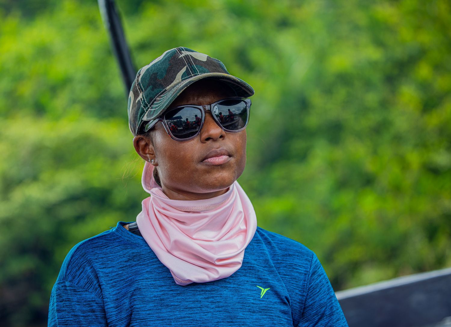 Person wearing camo cap, sunglasses, and pink neck gaiter in a lush green outdoor setting.