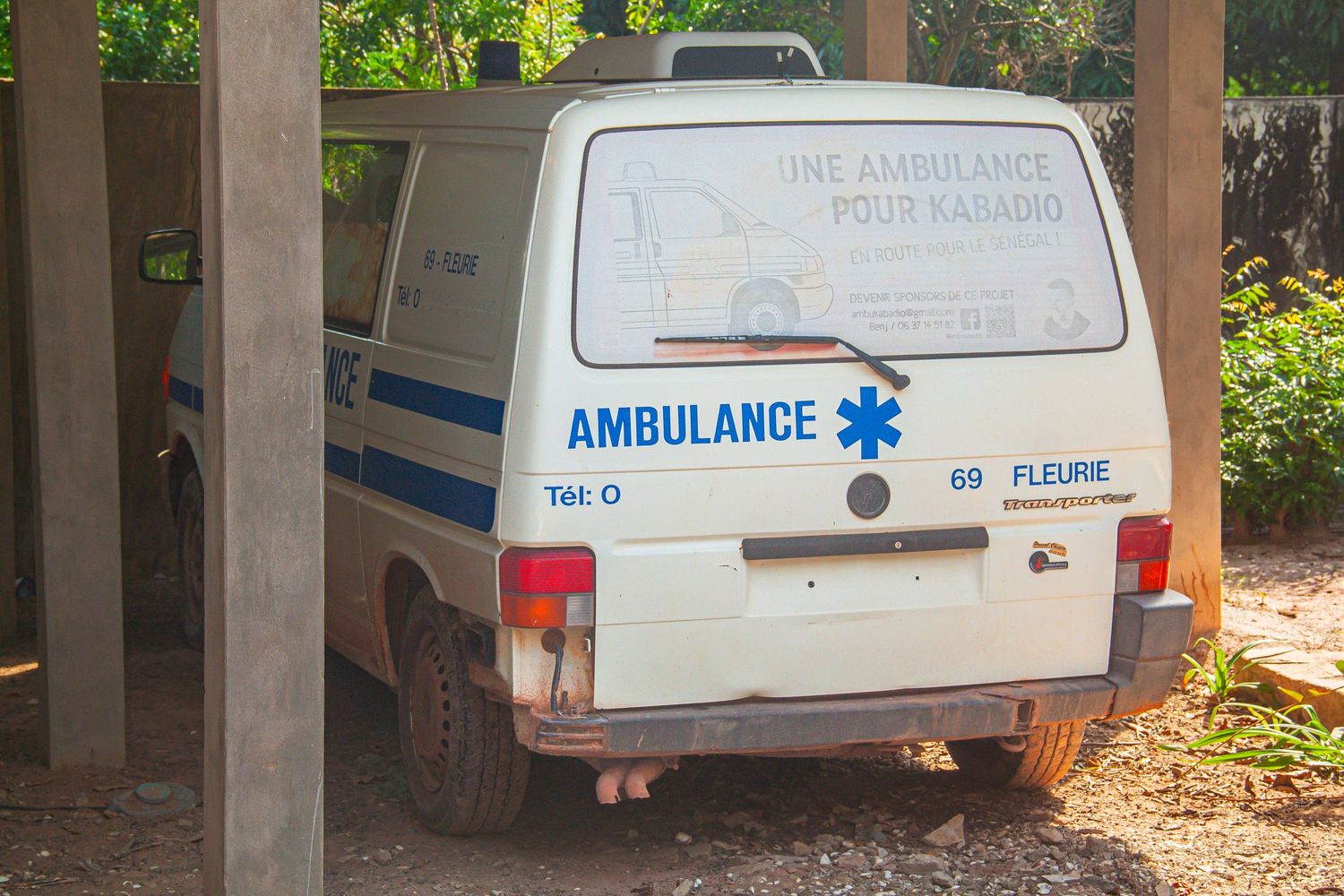 Old white ambulance van parked under a shelter in a rural African community setting.
