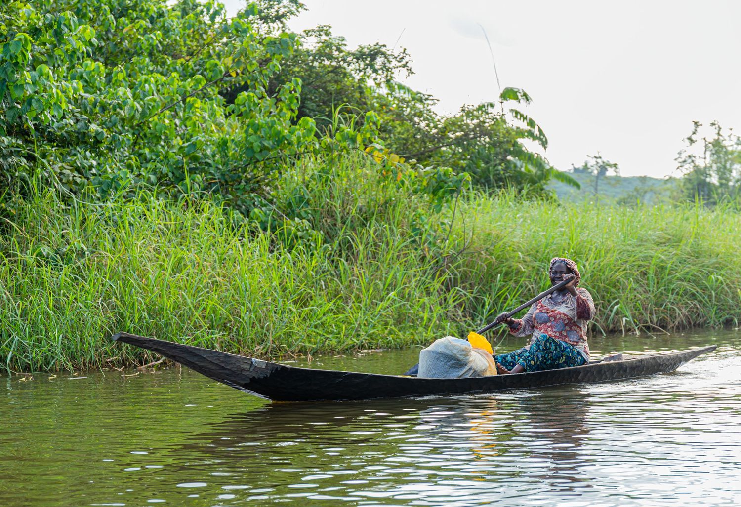 Woman paddling a traditional wooden canoe along a calm river surrounded by lush green vegetation in Africa.