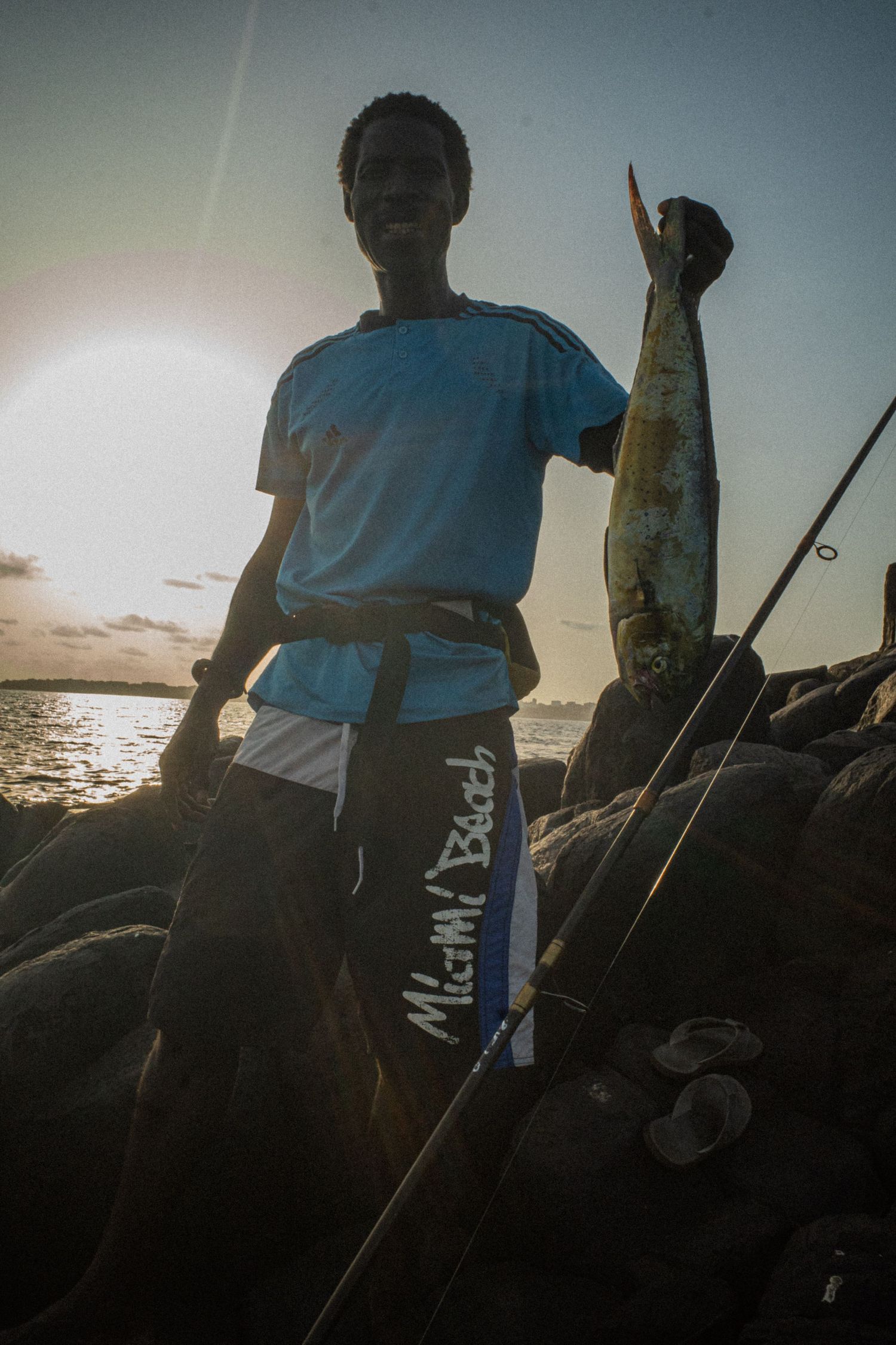 Young Senegalese fisherman holding a freshly caught fish on rocky shoreline at sunset.