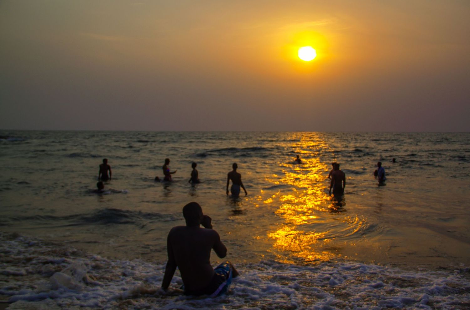 Silhouette of a person sitting on a beach at golden sunset while swimmers enjoy the ocean waves.