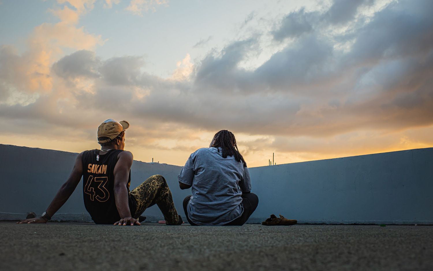 Two people sit side by side on a concrete surface watching a dramatic cloudy sunset together.