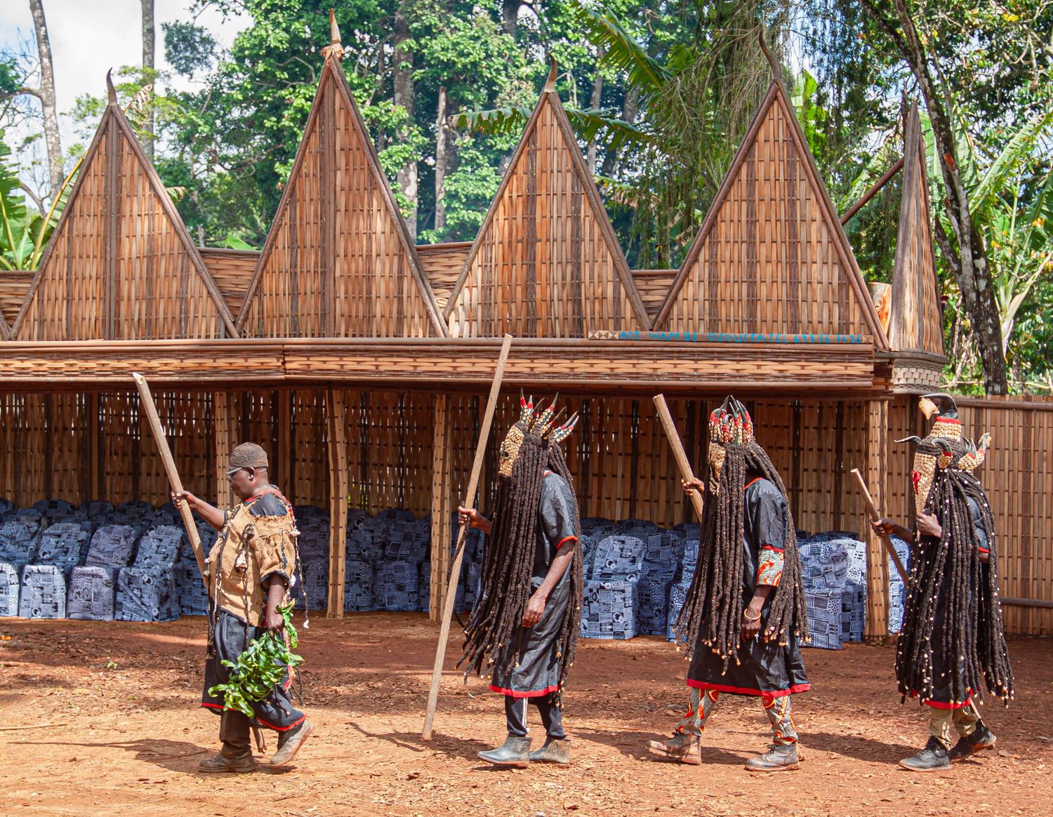 Four performers in traditional African ceremonial costumes with masks and staffs before a woven building.