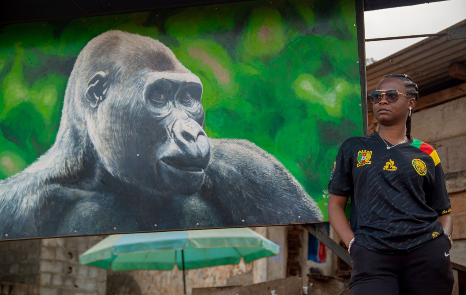 Man in Cameroon football jersey standing beside a large gorilla mural painted on a wall outdoors.