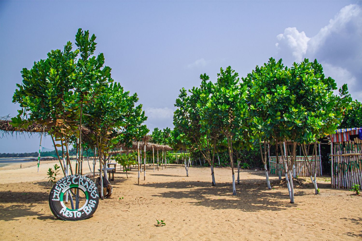 Lush green trees line a sandy beach with a rustic sign and wooden structures in a tropical setting.