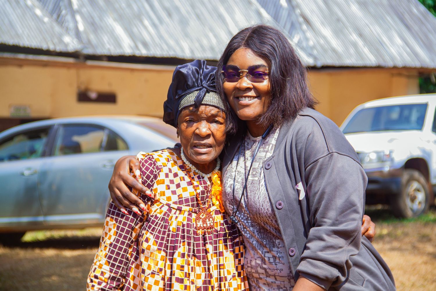 Two African women smiling and embracing outdoors, one in colorful traditional print dress.