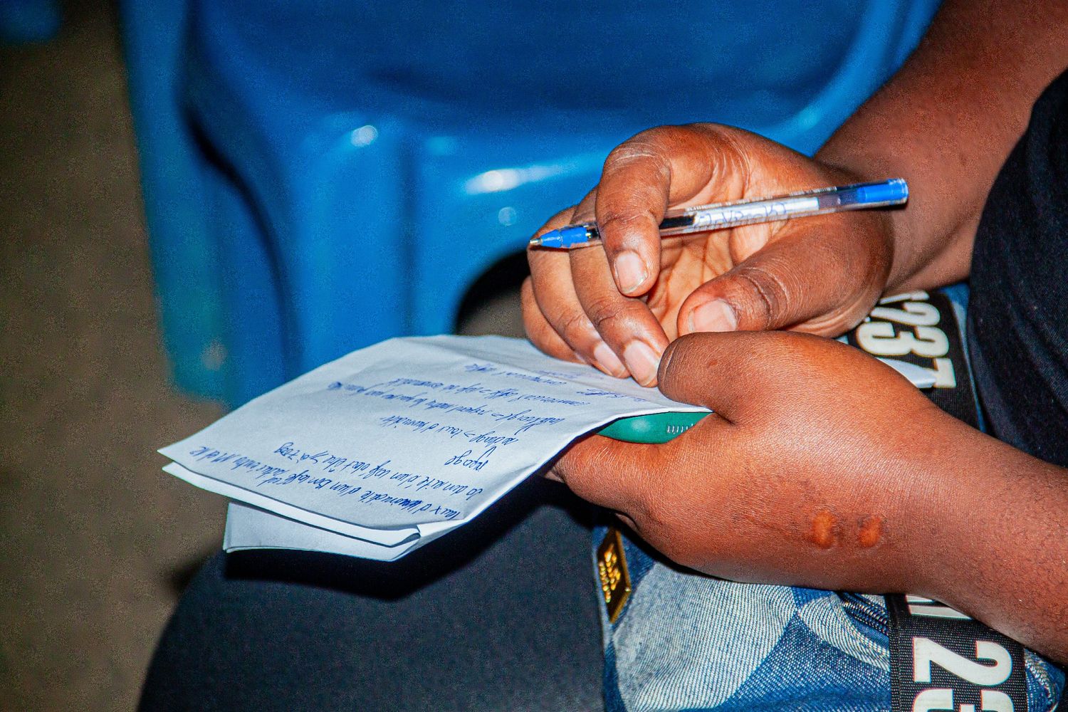 Close-up of hands writing notes on paper with a blue pen near a blue chair.