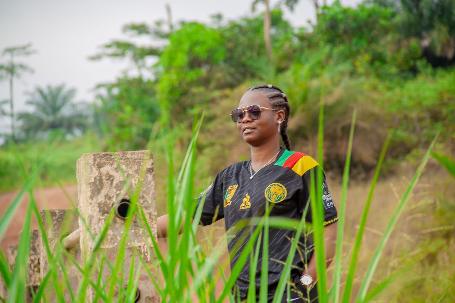 Person wearing Cameroon jersey and sunglasses standing in rural green landscape.