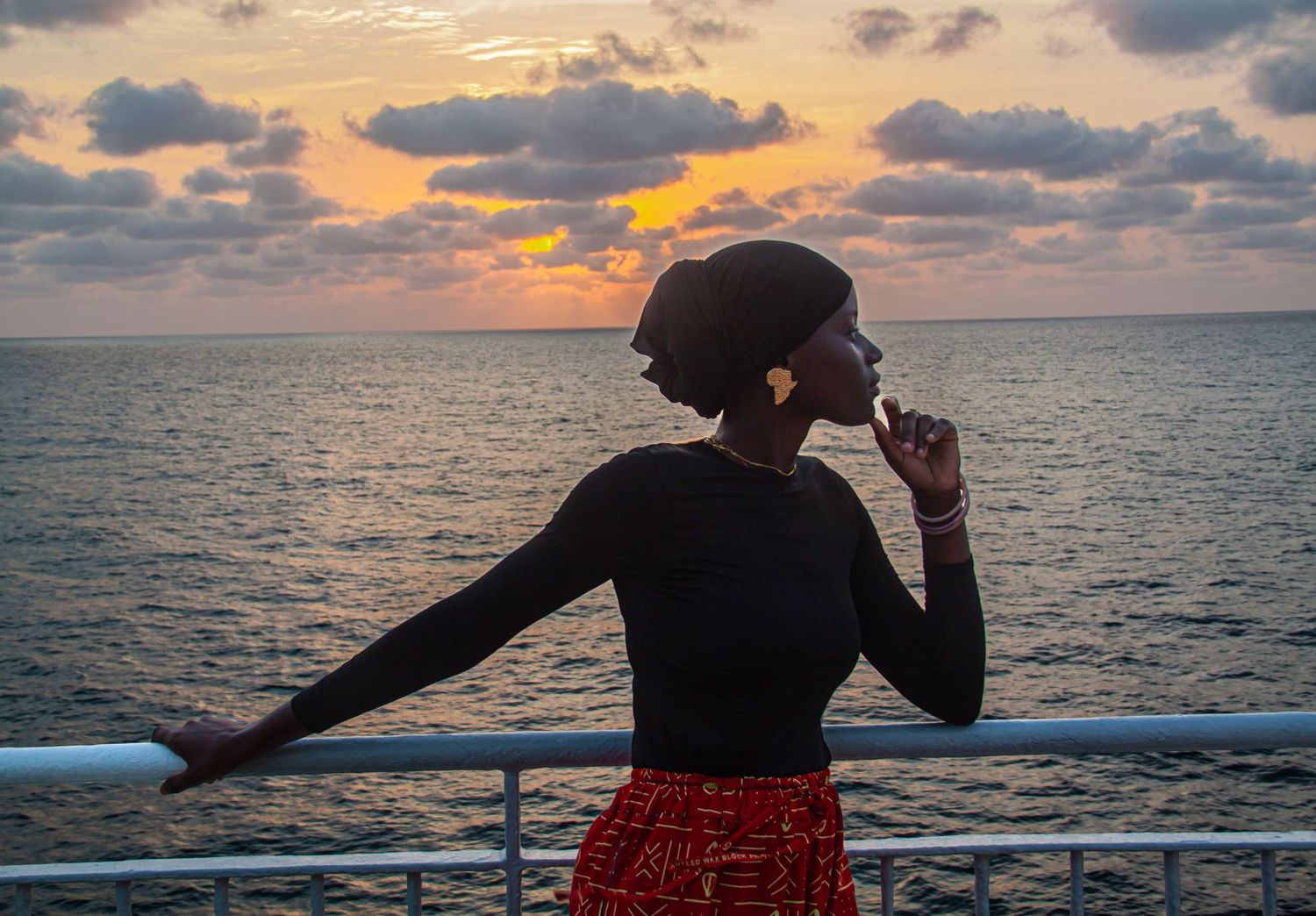 Elegant woman in black top and red skirt watching a stunning ocean sunset from a deck.