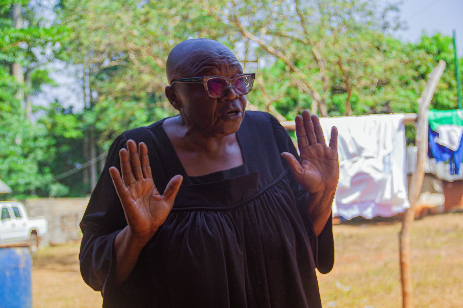 Bald African woman in black dress with pink glasses raising both hands outdoors.
