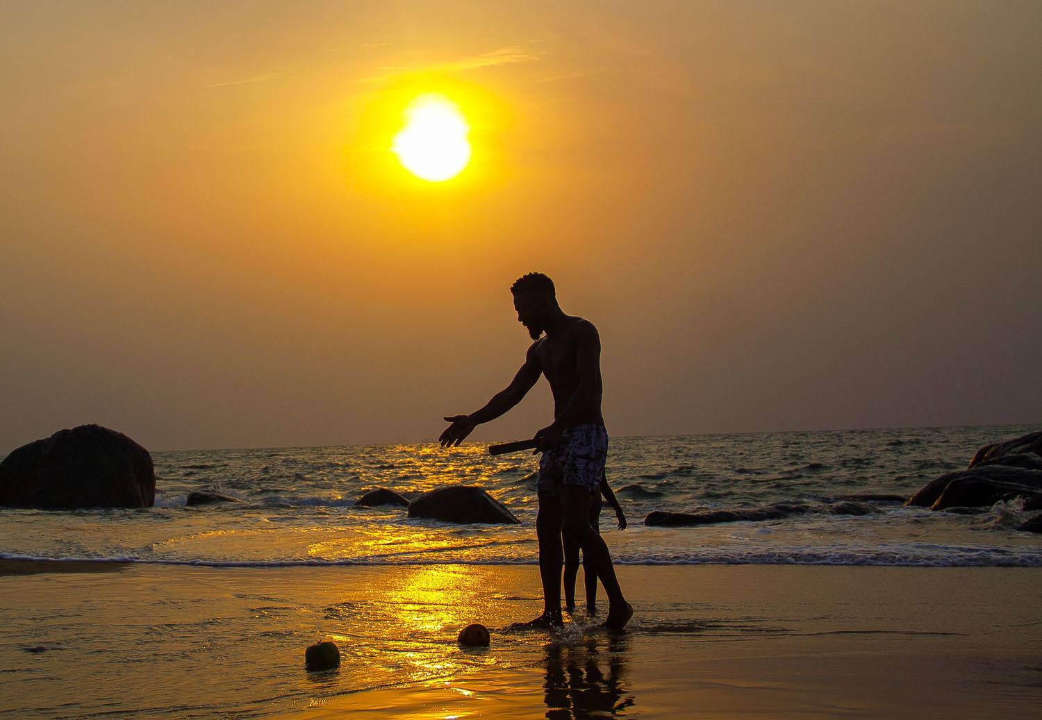 Silhouette of two people playing on a reflective beach at golden sunset with rocks in the ocean.