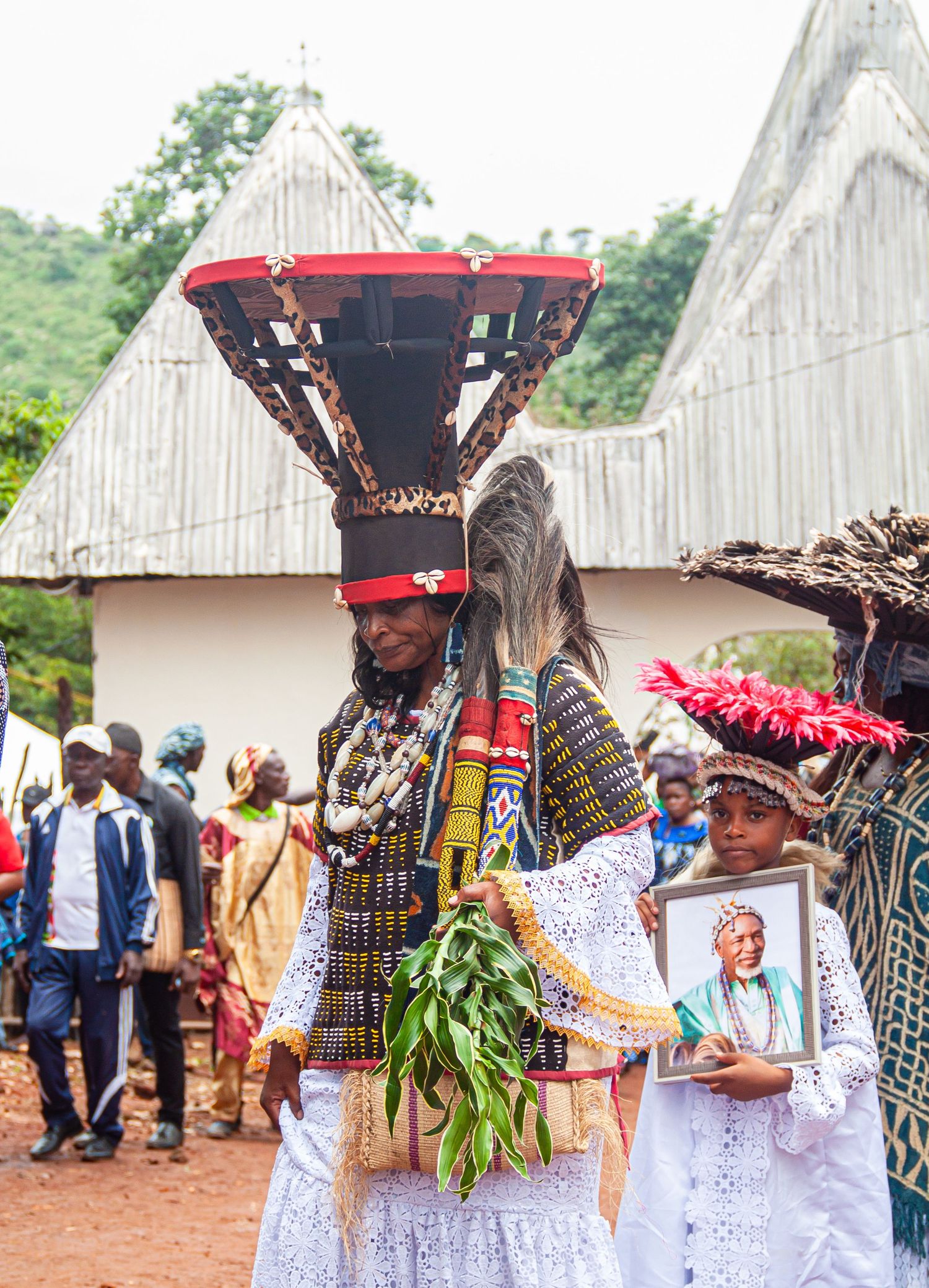Person in elaborate traditional African ceremonial costume with tall headdress and child holding portrait at festival.