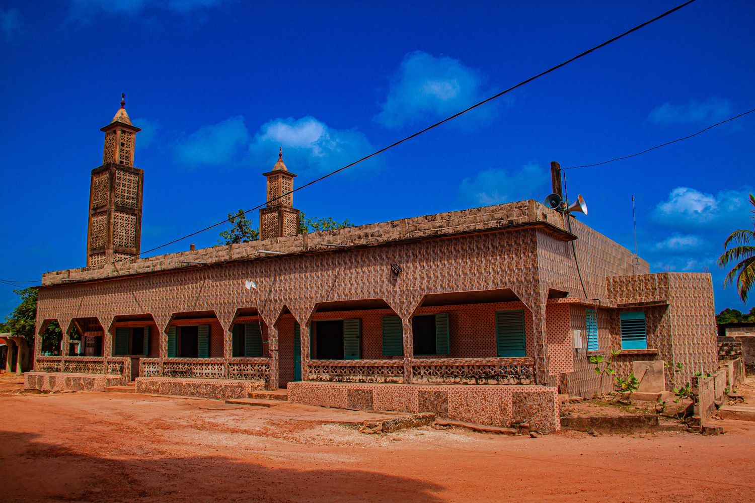 Traditional West African mosque with decorative mud-brick architecture, twin minarets, and arched colonnade under vivid blue sky.