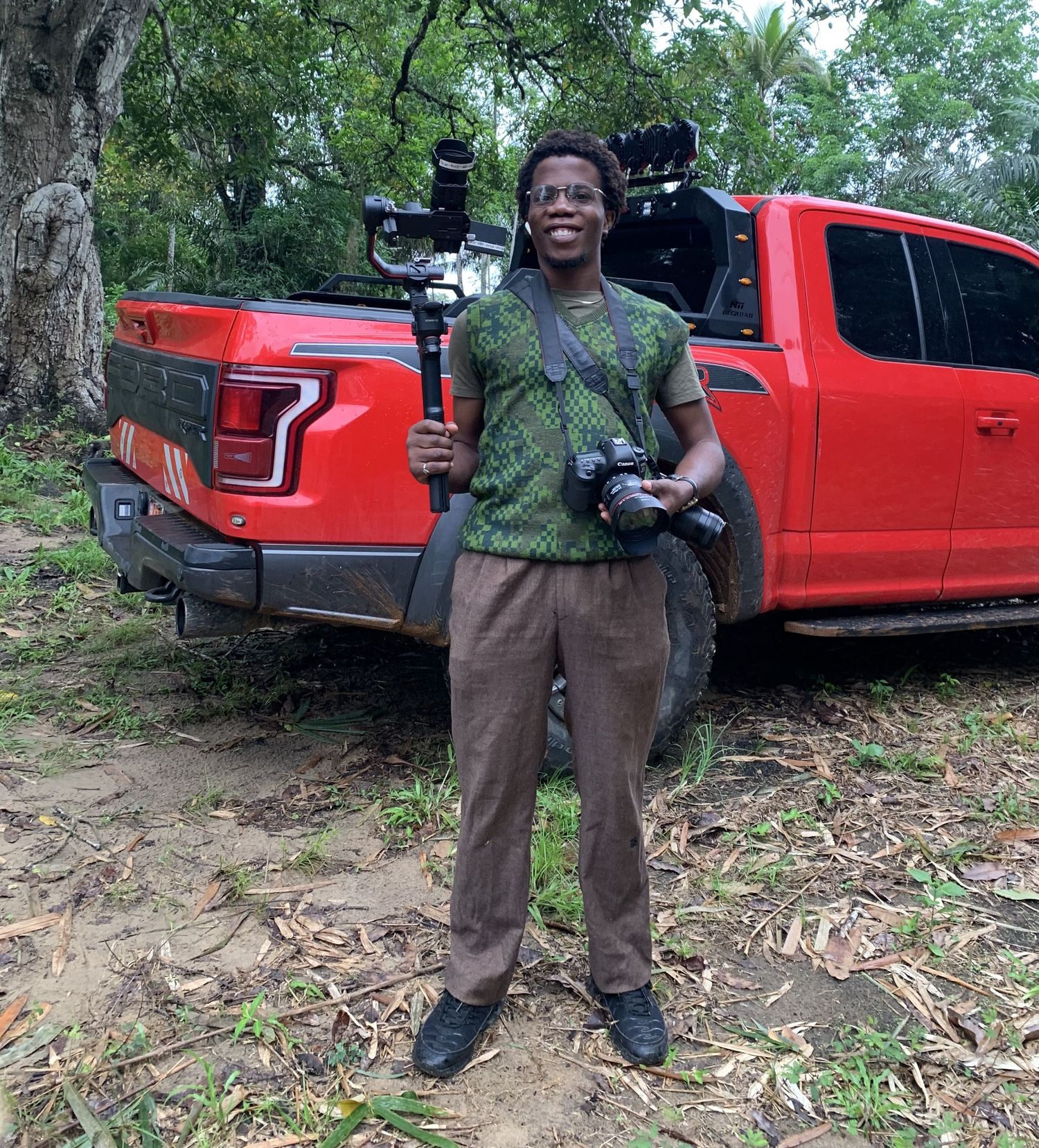 African photographer standing beside a red pickup truck holding a camera gimbal stabilizer outdoors.