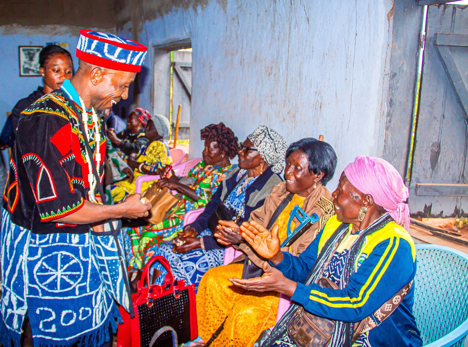 Traditional African ceremony with elder in colorful attire greeting seated women in vibrant cultural dress.