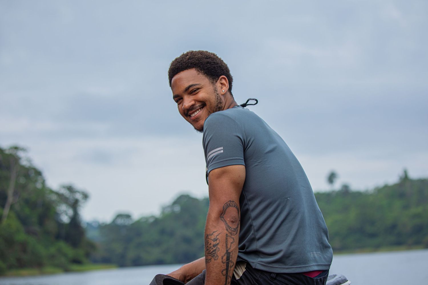 A smiling man with a tattoo sits on a boat near a lush tropical river.