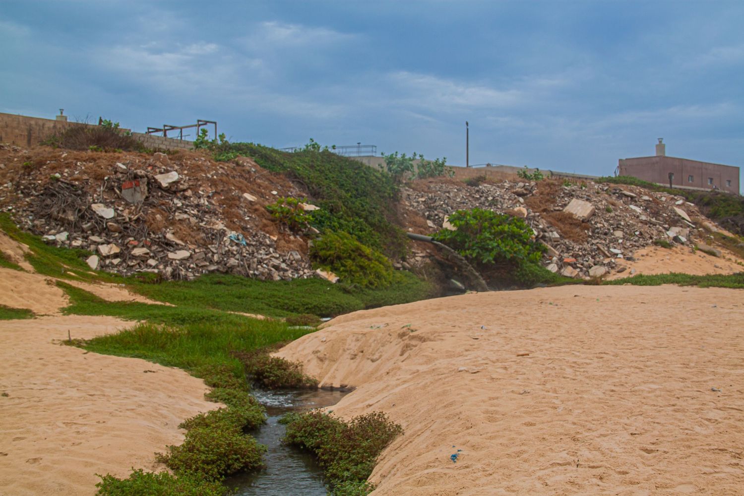 A sandy beach with a small stream, rocky debris, and green vegetation under an overcast sky.