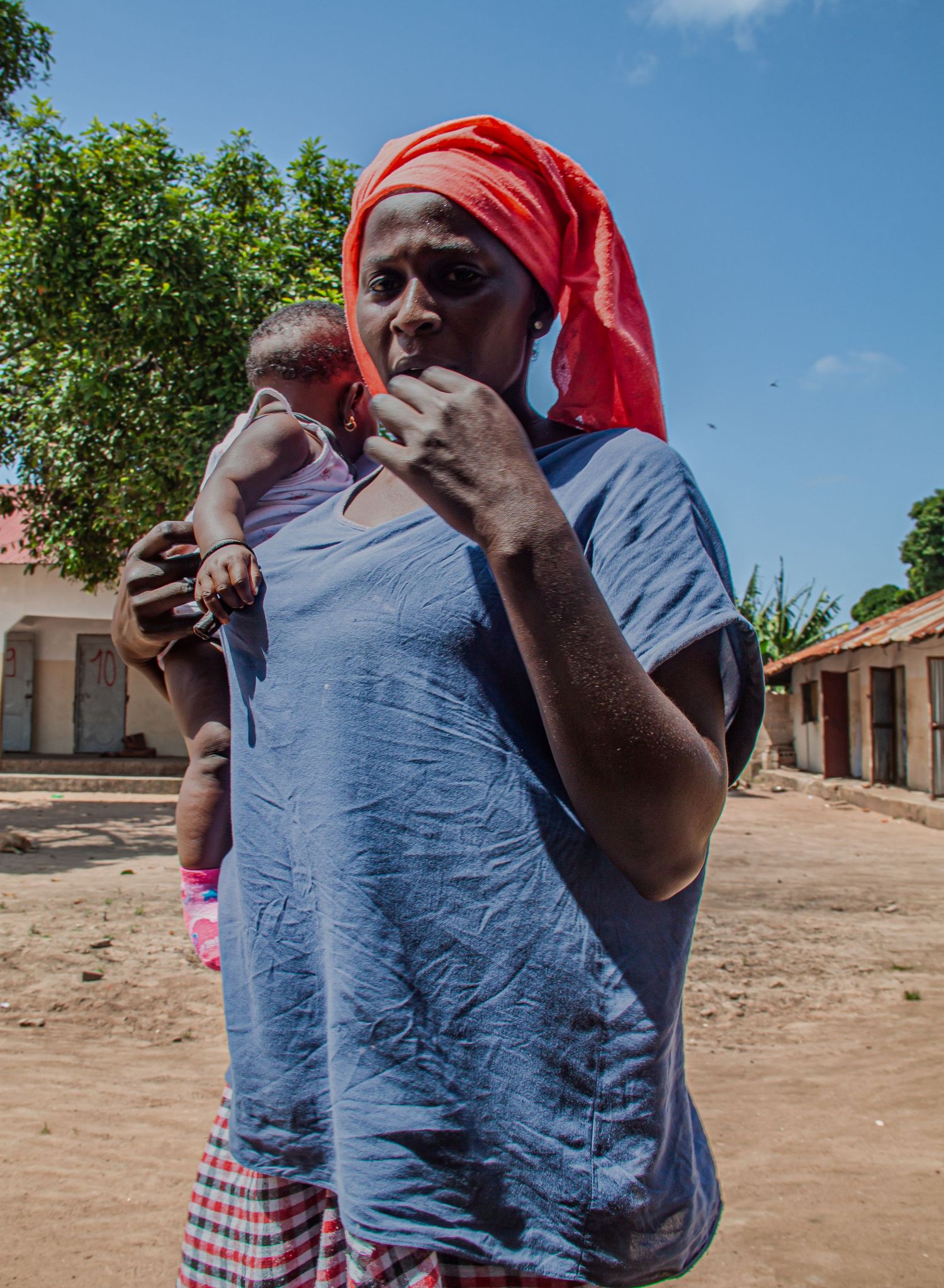 Senegalese mother in blue dress and red headwrap carrying her baby outdoors on a sunny day.