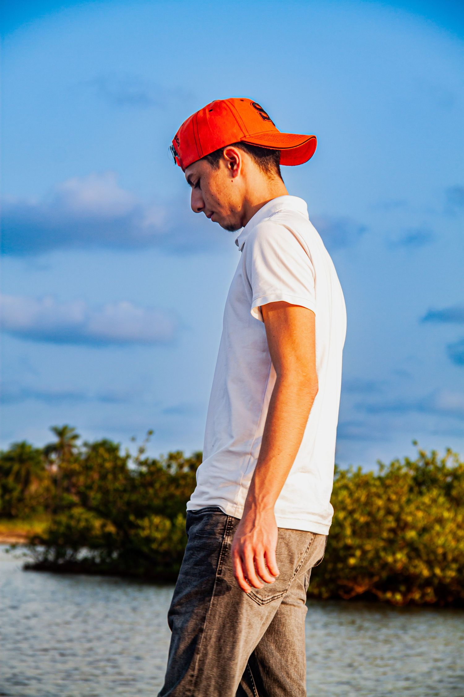 Young man in red cap and white polo shirt standing outdoors near water with blue sky background.