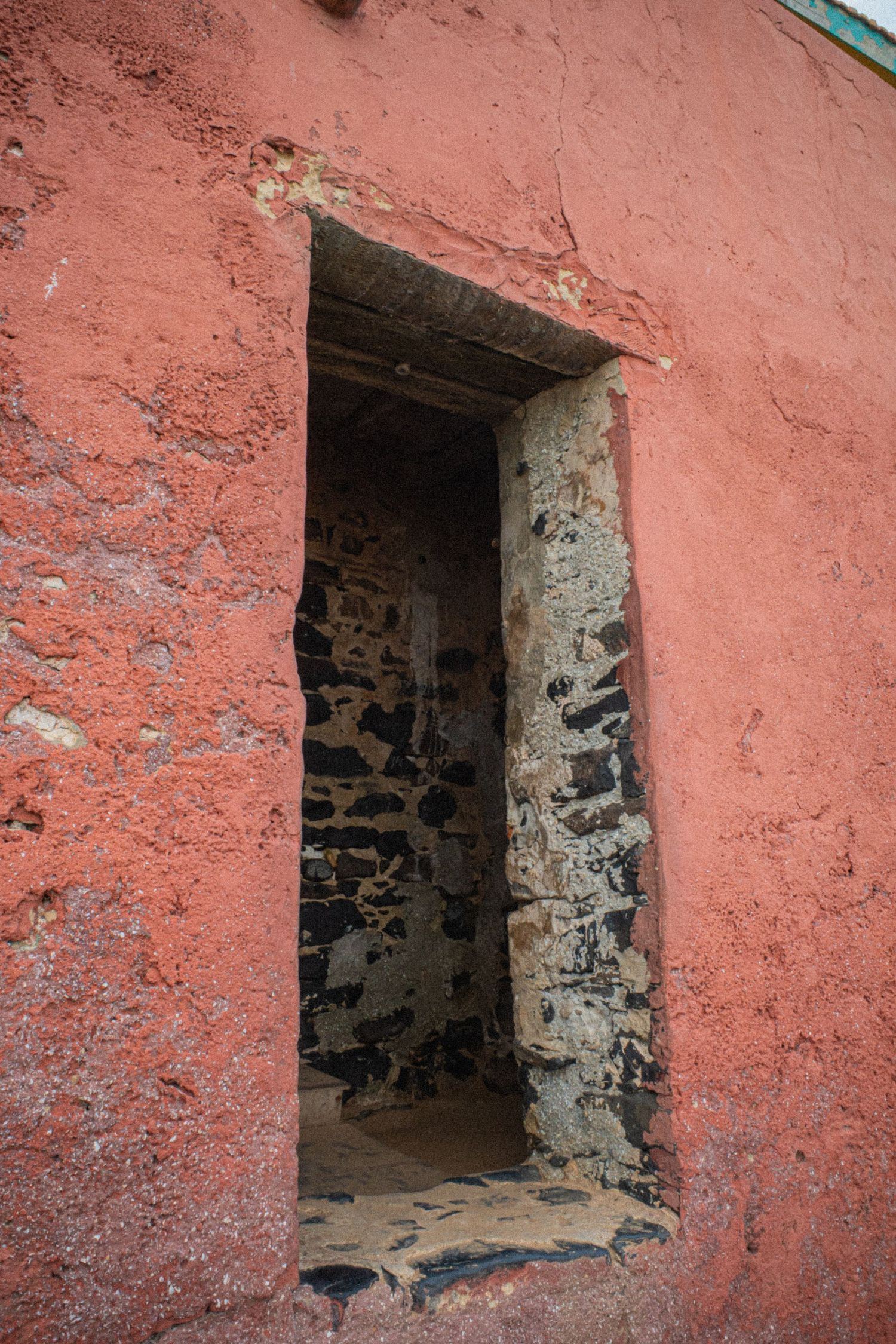 A weathered red-painted stone wall with a small rectangular doorway revealing dark interior stones.