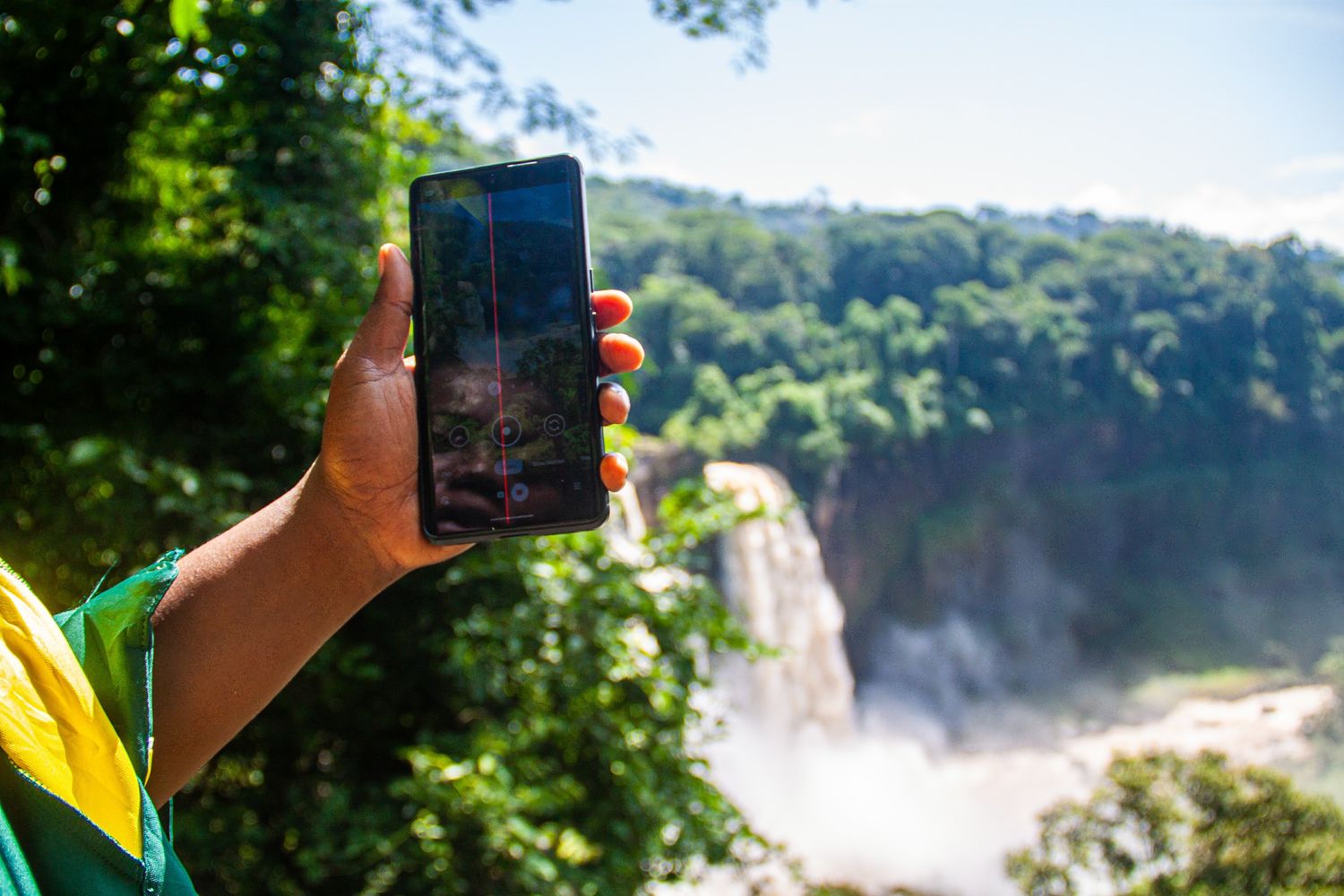 Hand holding smartphone photographing a scenic tropical waterfall surrounded by lush greenery.