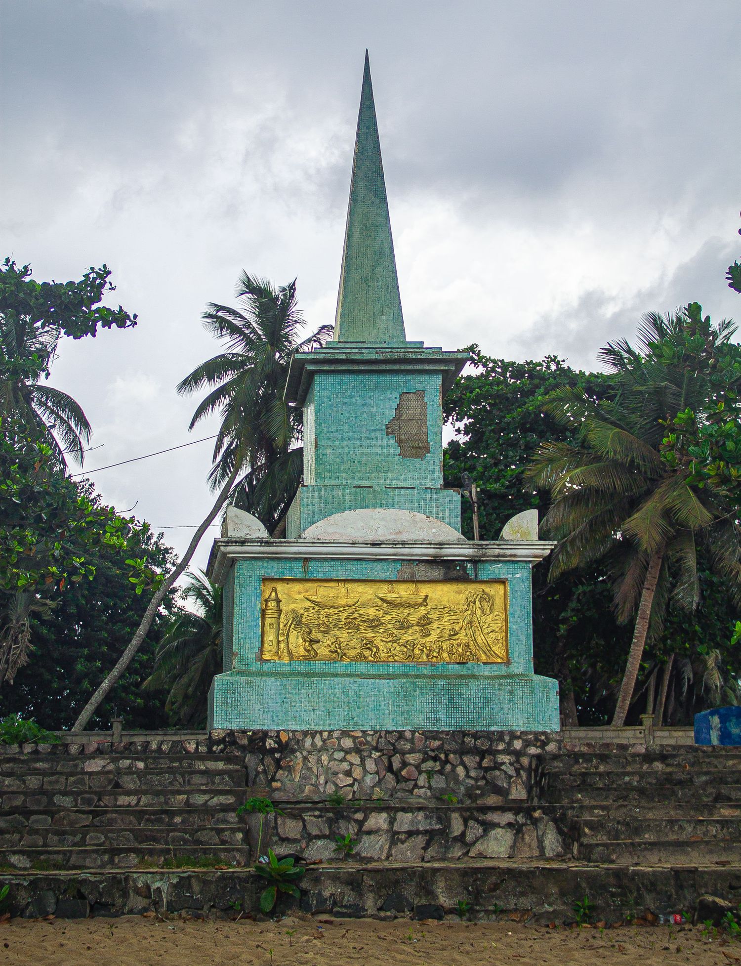 Weathered teal obelisk monument with gold relief plaque surrounded by palm trees under cloudy sky.