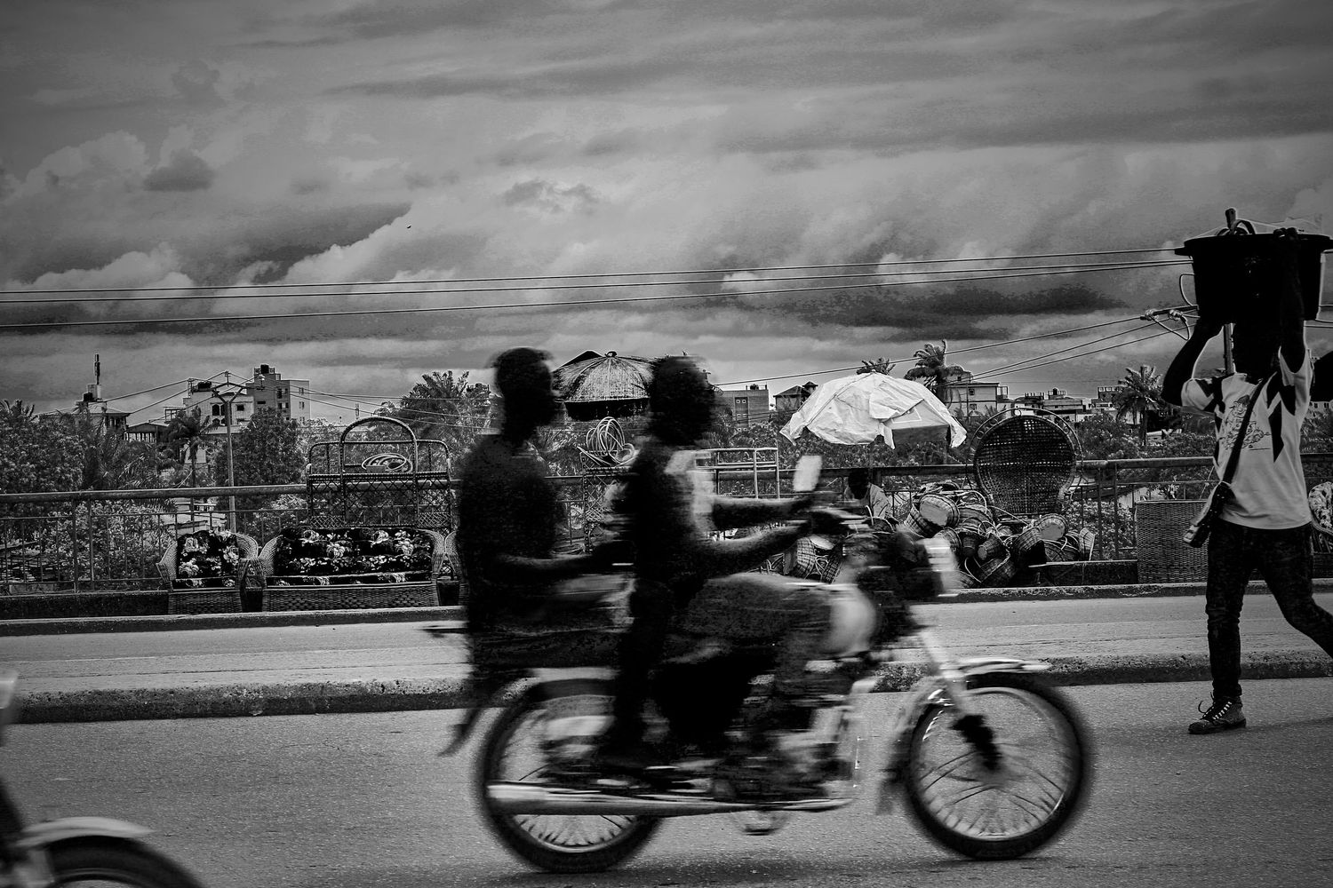 Black and white photo of people riding a motorcycle on a busy African road under dramatic clouds.