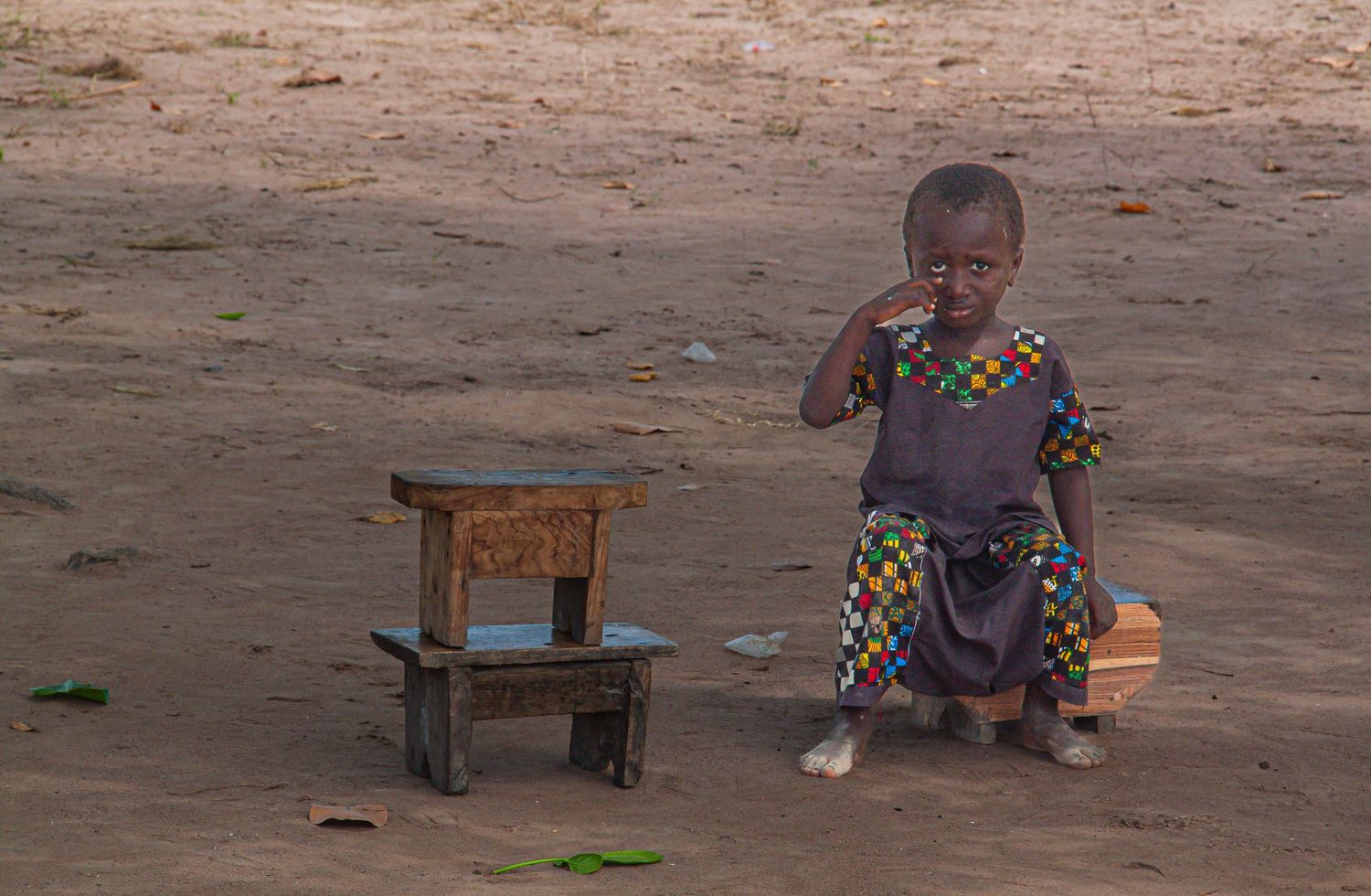 Young African child standing next to a small wooden stool on sandy ground in a rural village.