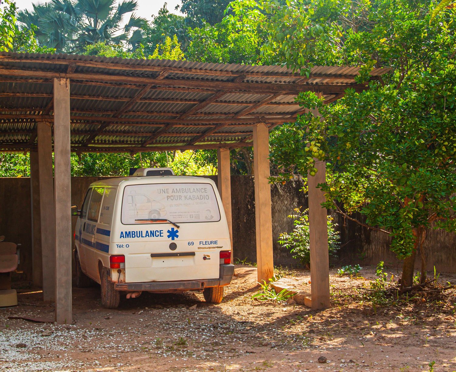 Old white ambulance van parked under a rustic wooden canopy shelter surrounded by trees.