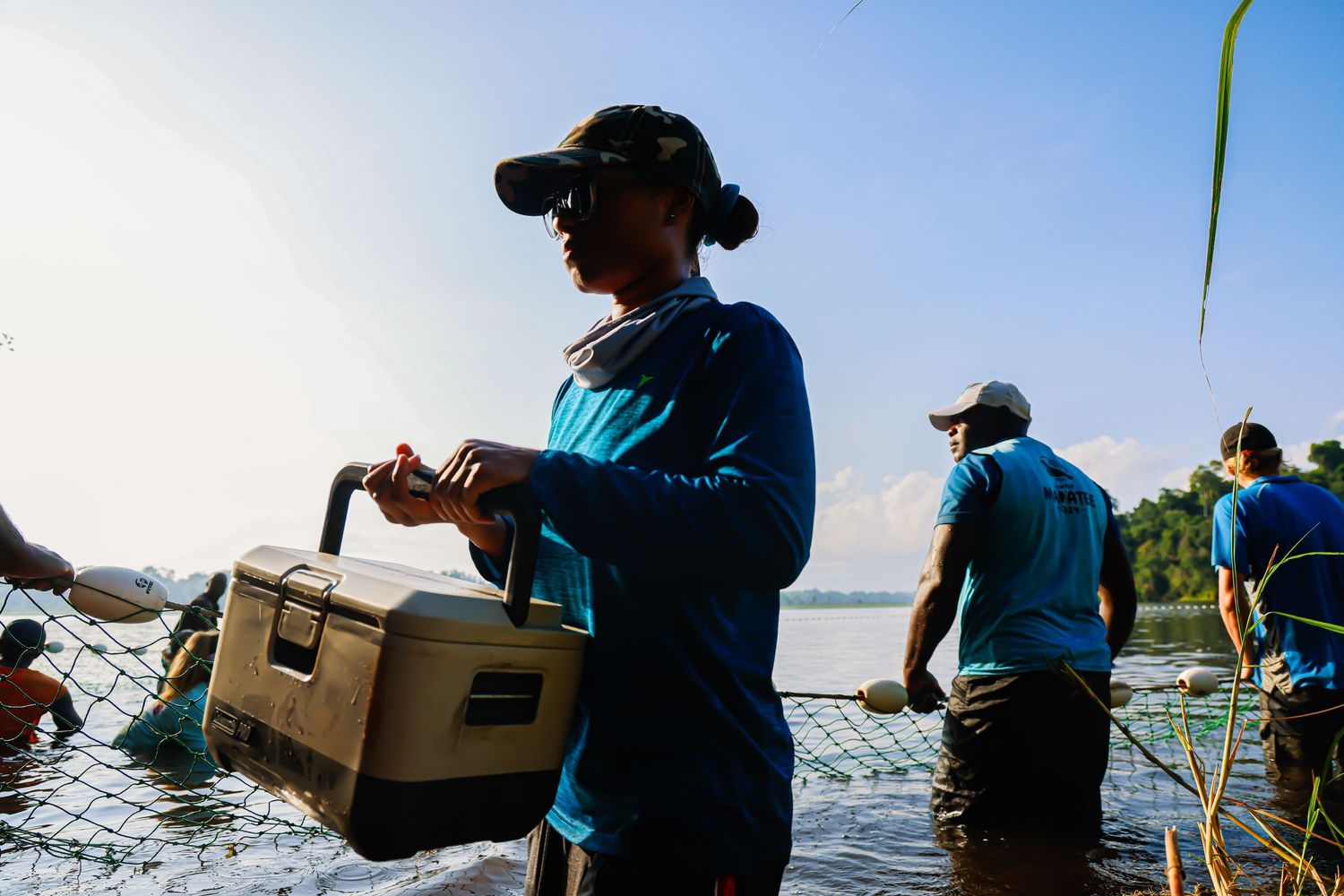 A person in a blue shirt and cap holds a cooler while standing in water during a group fishing activity at sunset.
