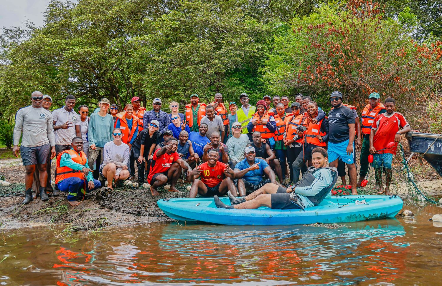 A large diverse group of conservation volunteers poses together on a tropical riverbank with kayaks and boats.