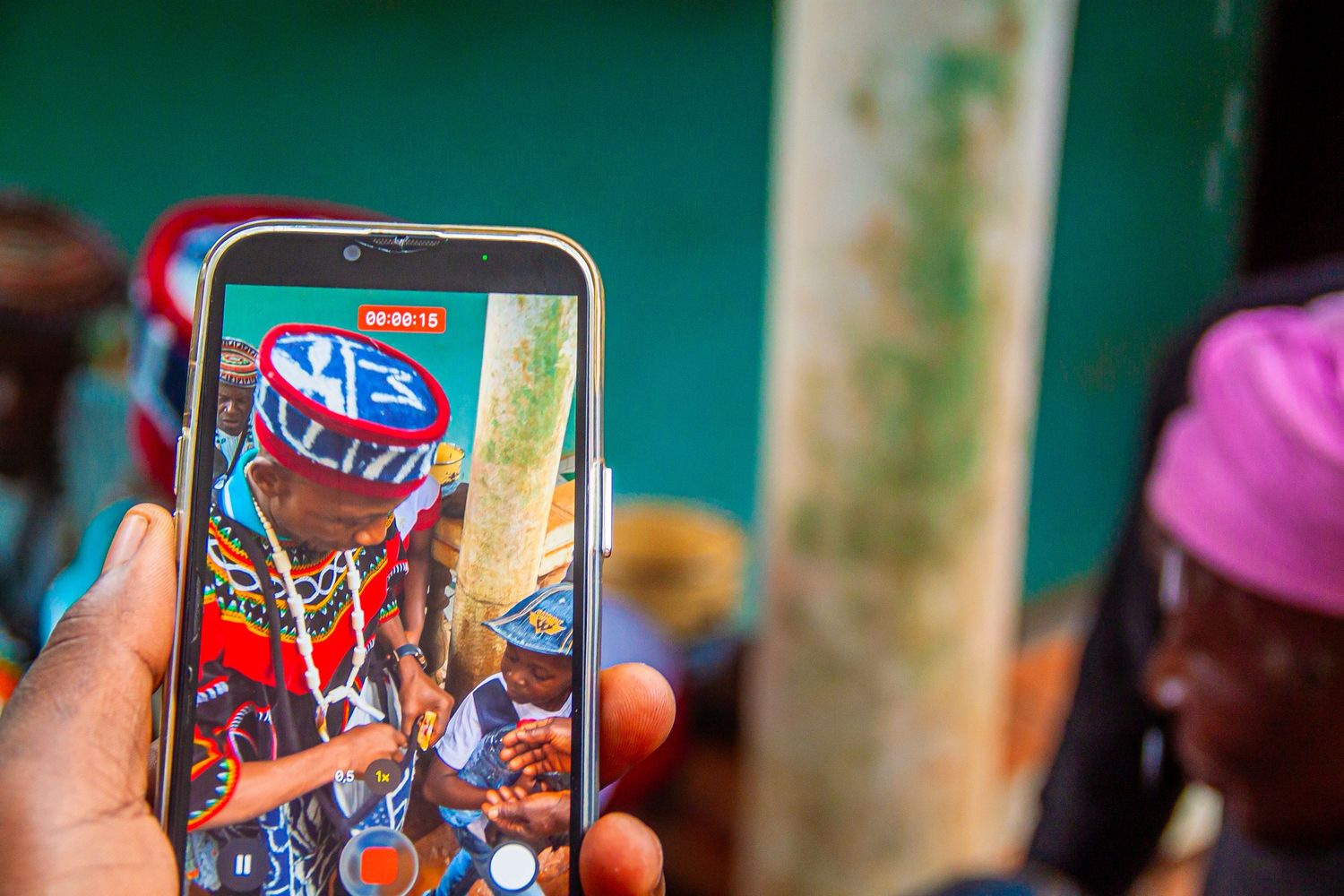 Hand holding smartphone displaying photo of person in colorful traditional African attire and beaded headwear at cultural event.
