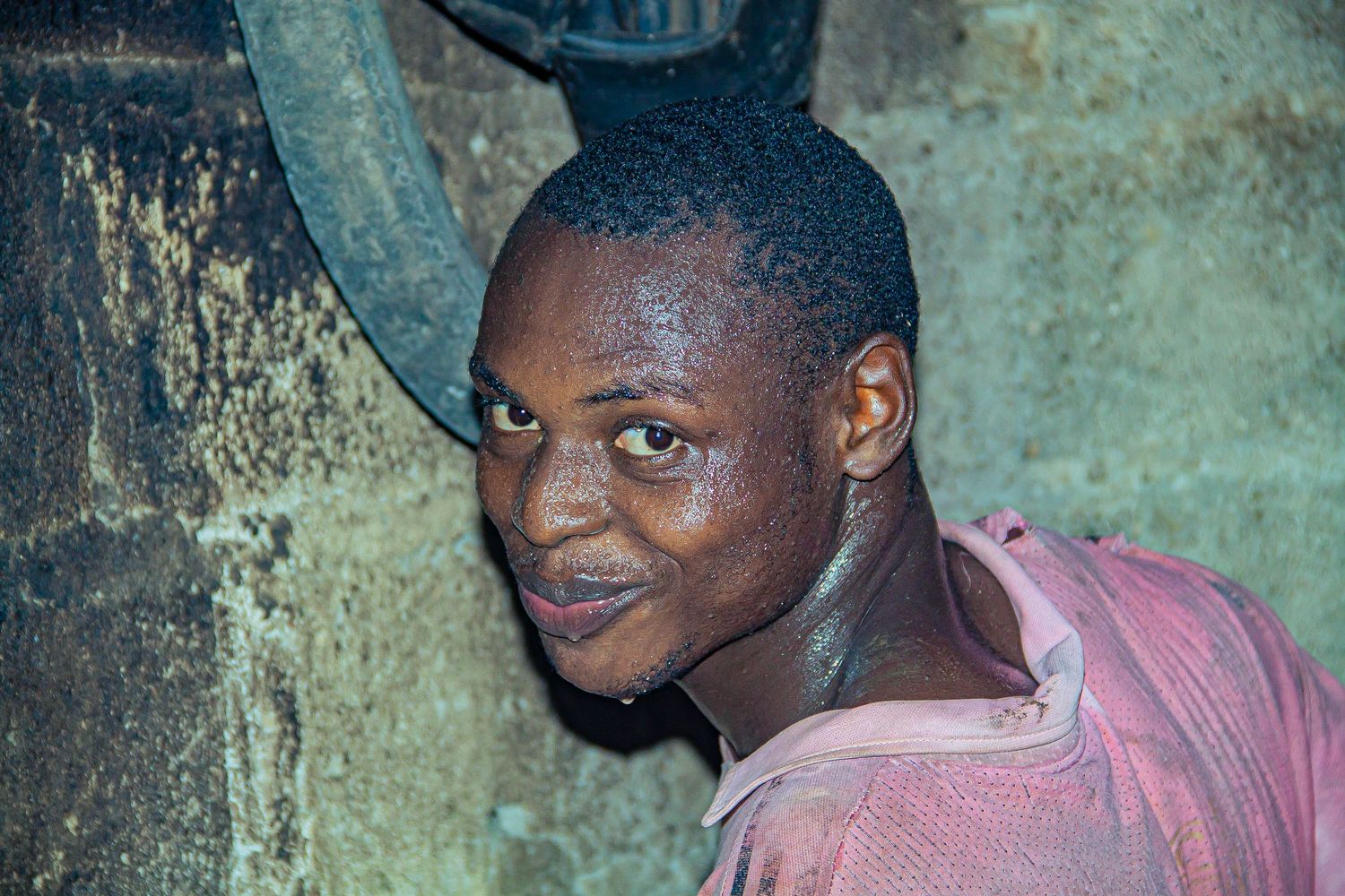 A young African man in a pink shirt smiles near industrial equipment in dim lighting.