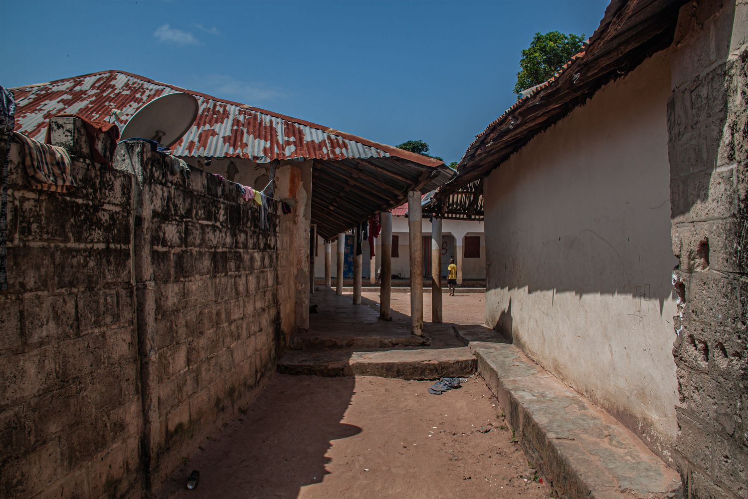 Narrow dusty alleyway between weathered concrete walls and rustic buildings in a West African village.