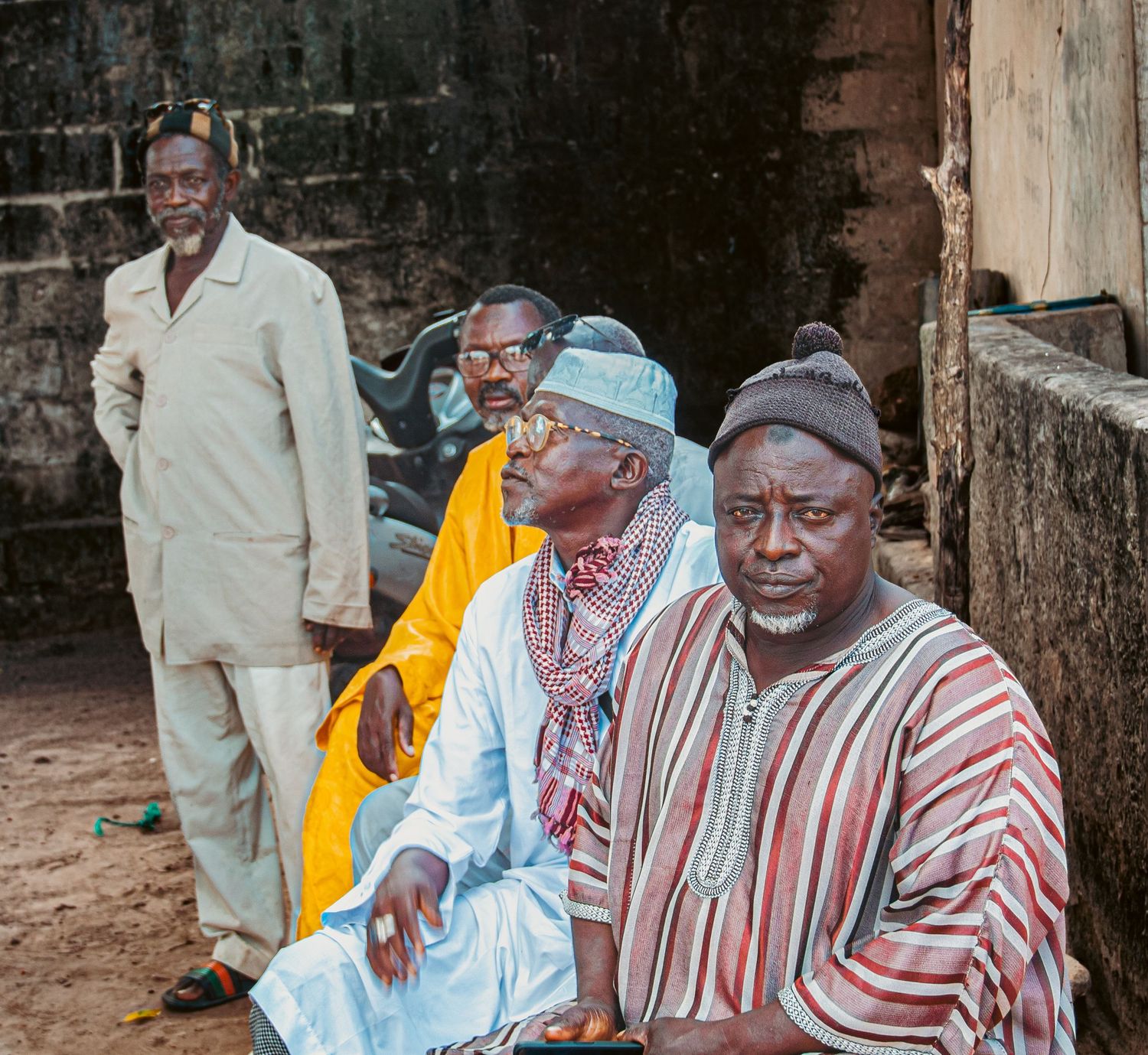 Group of elder African men in traditional robes sitting and standing against an old stone wall.