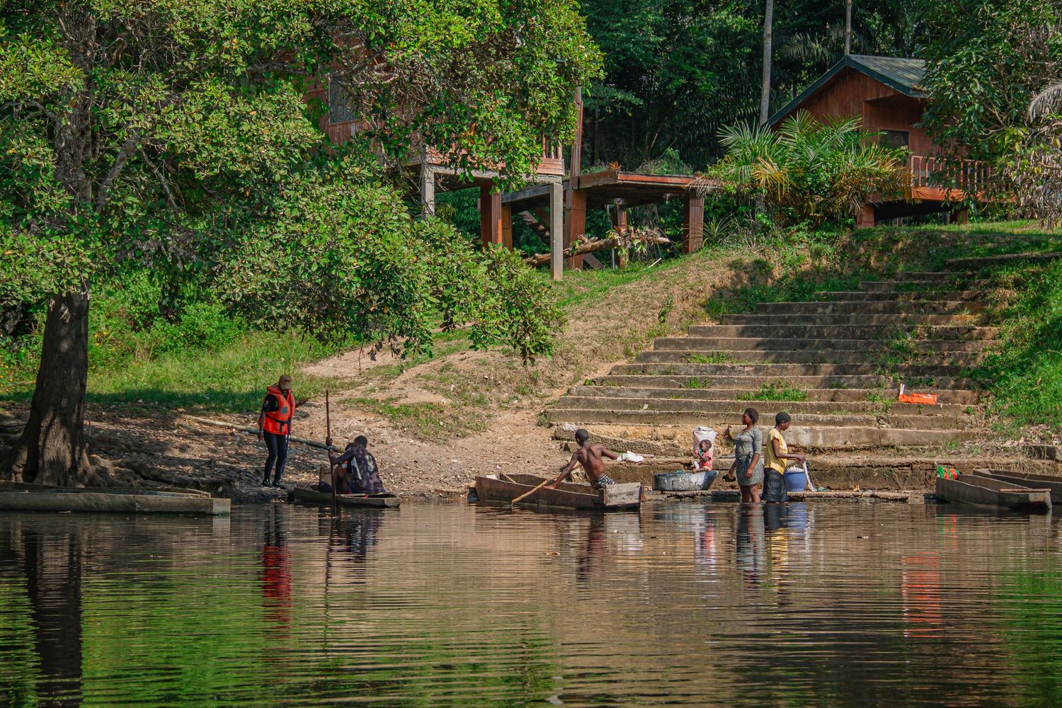 Group of people washing clothes and boats on a calm river bank surrounded by lush tropical forest.