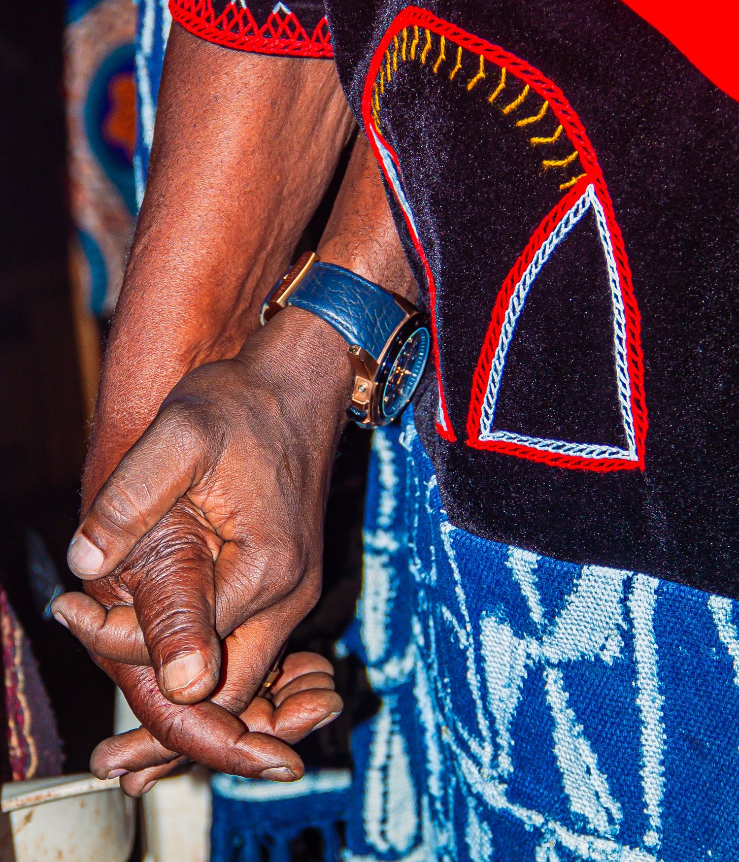 Close-up of two African people holding hands, wearing traditional embroidered cultural clothing.