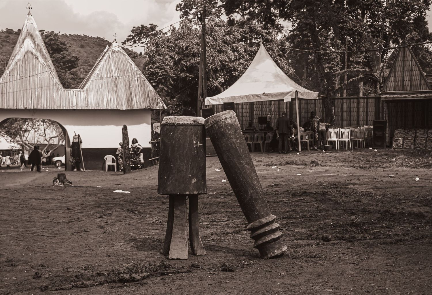 Black and white photo of traditional wooden drums standing in an African ceremonial grounds.