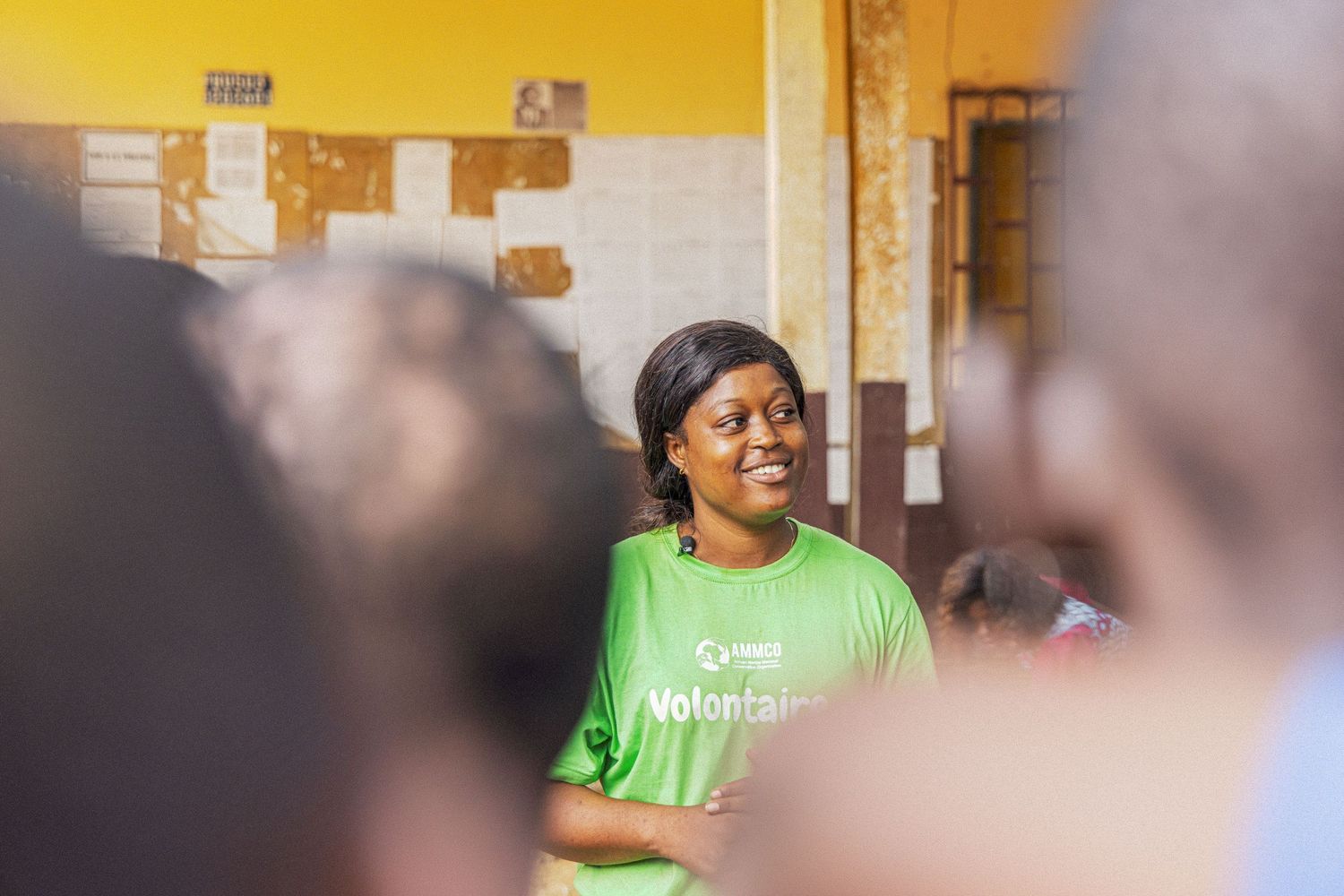 Woman in green volunteer t-shirt speaking to a group inside a community classroom.